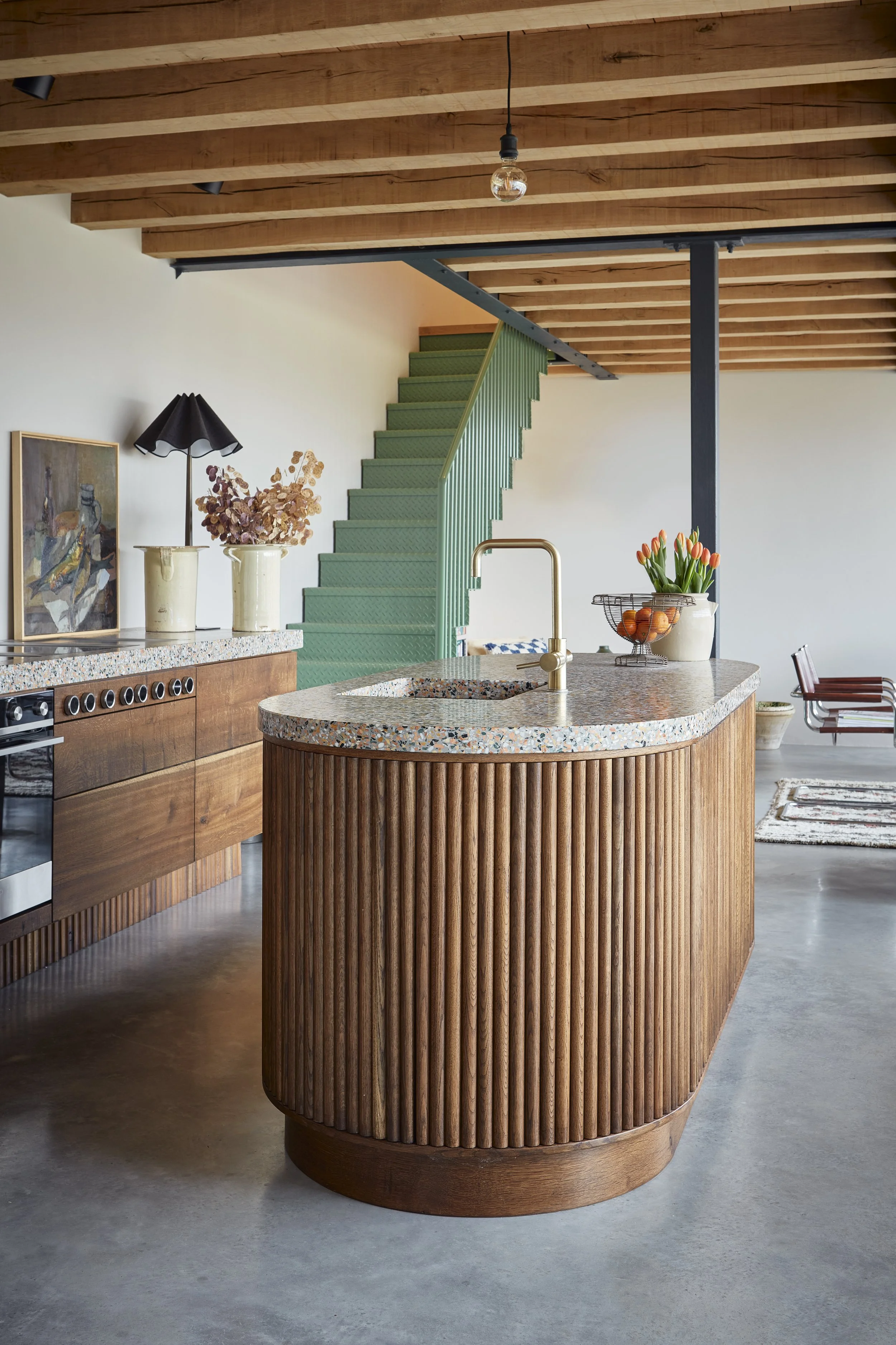 Interior view of a modern kitchen with a curved wooden island, terrazzo countertop, and brass faucet. There are potted plants, a table lamp, and artwork on the counter. A staircase with green carpeting and a wooden ceiling are visible in the backgrou