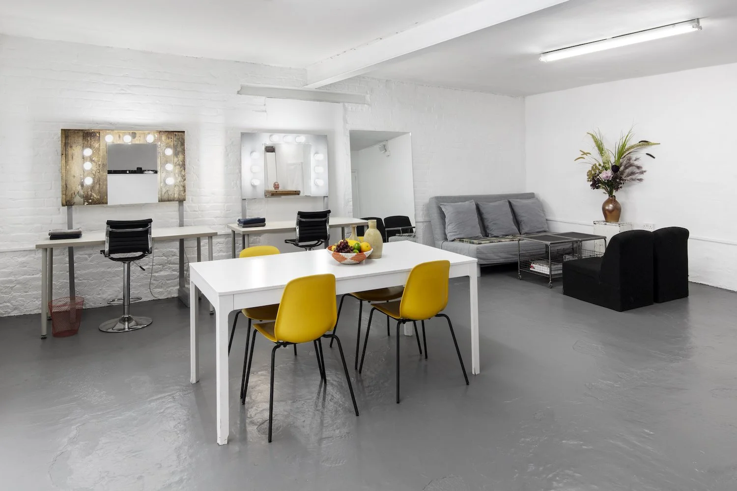 A spacious, minimalist room with white brick walls, a gray floor, and a white ceiling. It features a white table with four yellow chairs, a second table with black chairs and lighted mirrors, a gray couch with pillows, a flower vase with tall flowers
