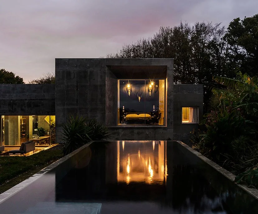 Modern house at dusk with illuminated bedroom visible through large window, a reflecting pool in the foreground, and lush trees surrounding the property.