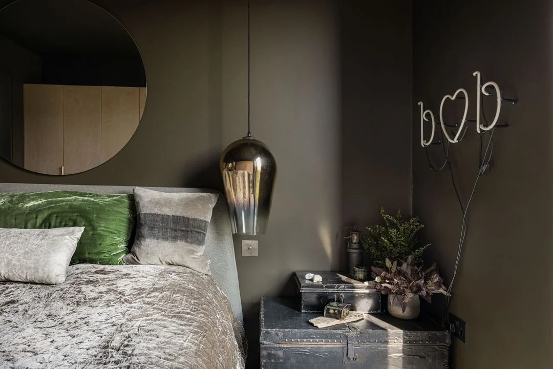 A bedroom with dark gray walls, a bed with a gray and green velvet pillow, a large round mirror on the wall, a metallic pendant light hanging above a black nightstand, which holds a potted plant, a small black box, and some decorative items. There is