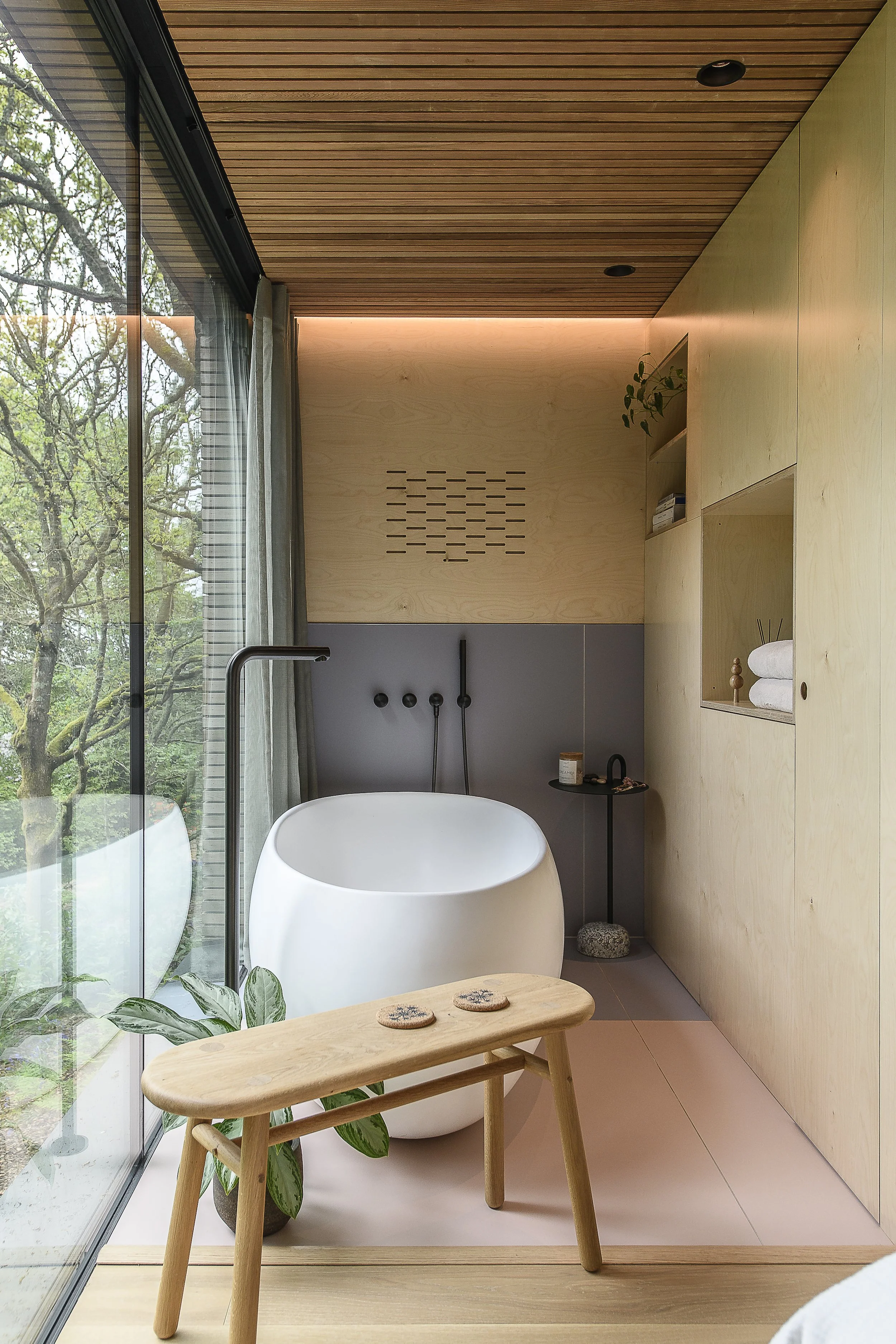Modern bathroom with a freestanding white bathtub, wooden bench with decorative stones, large glass window overlooking trees, and built-in wooden shelves with towels and decor.