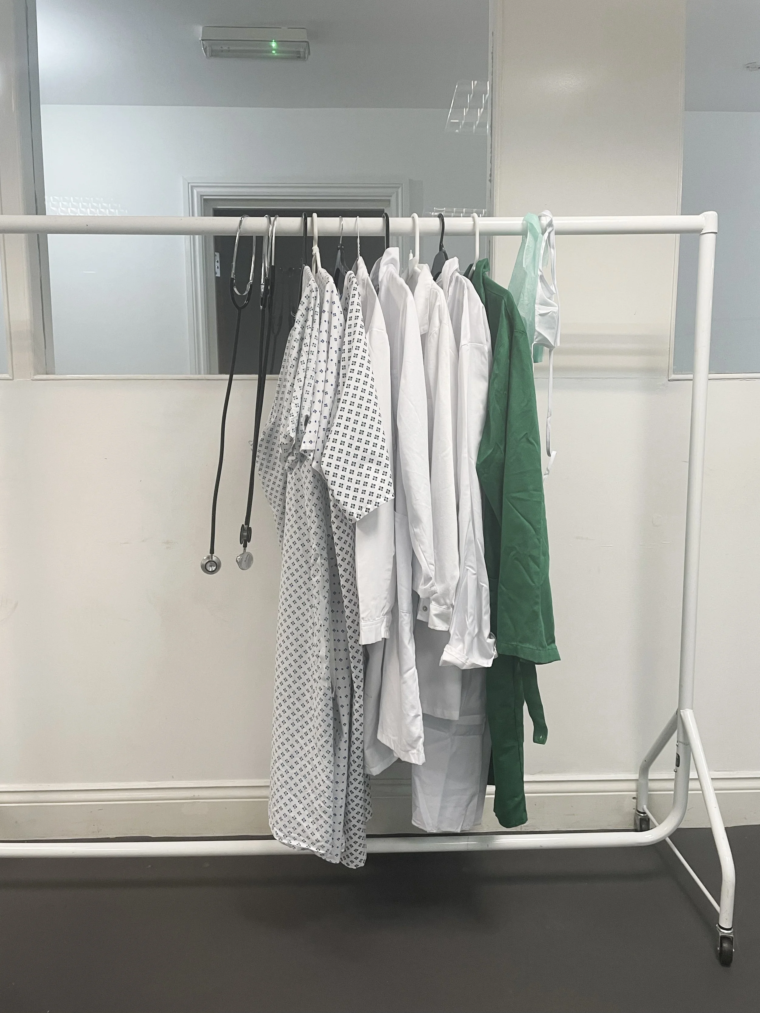 Clothes hanging on a white metal clothing rack, including patterned and solid-colored medical scrubs and lab coats, with a stethoscope hanging on the rack.