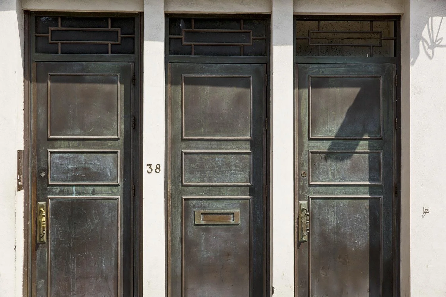 Three weathered metal doors with rectangular panels, two equipped with handles, and a white wall with the house number 38 to the left of the middle door.