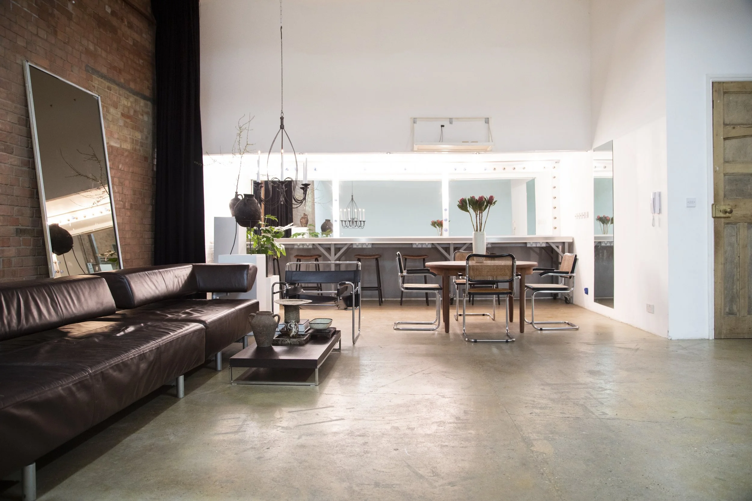 Interior of a spacious modern living area with a brown leather sofa, a large mirror, a coffee table with decorative items, and a dining area with a wooden table and chairs, featuring a bright, airy atmosphere with natural light.