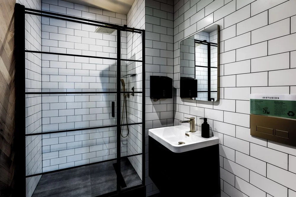 Modern bathroom with walk-in shower enclosed by black-framed glass, white subway tiles on walls, a small mirror above a black vanity with a white sink, a black soap dispenser, two black wall-mounted dispensers, and a partially visible wooden wall on 