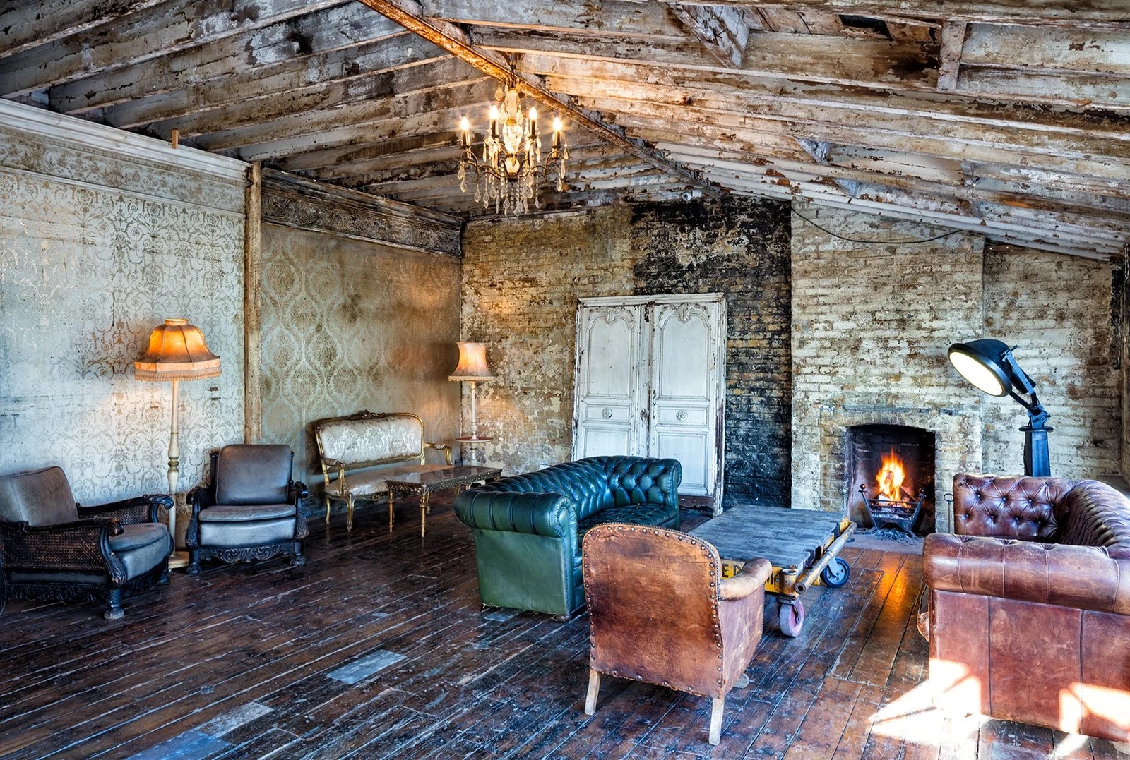 Interior of a rustic living room with exposed brick walls, a fireplace with a fire, vintage furniture including leather and fabric armchairs, a chandelier hanging from a sloped wooden ceiling, and a floor lamp with a spotlight.