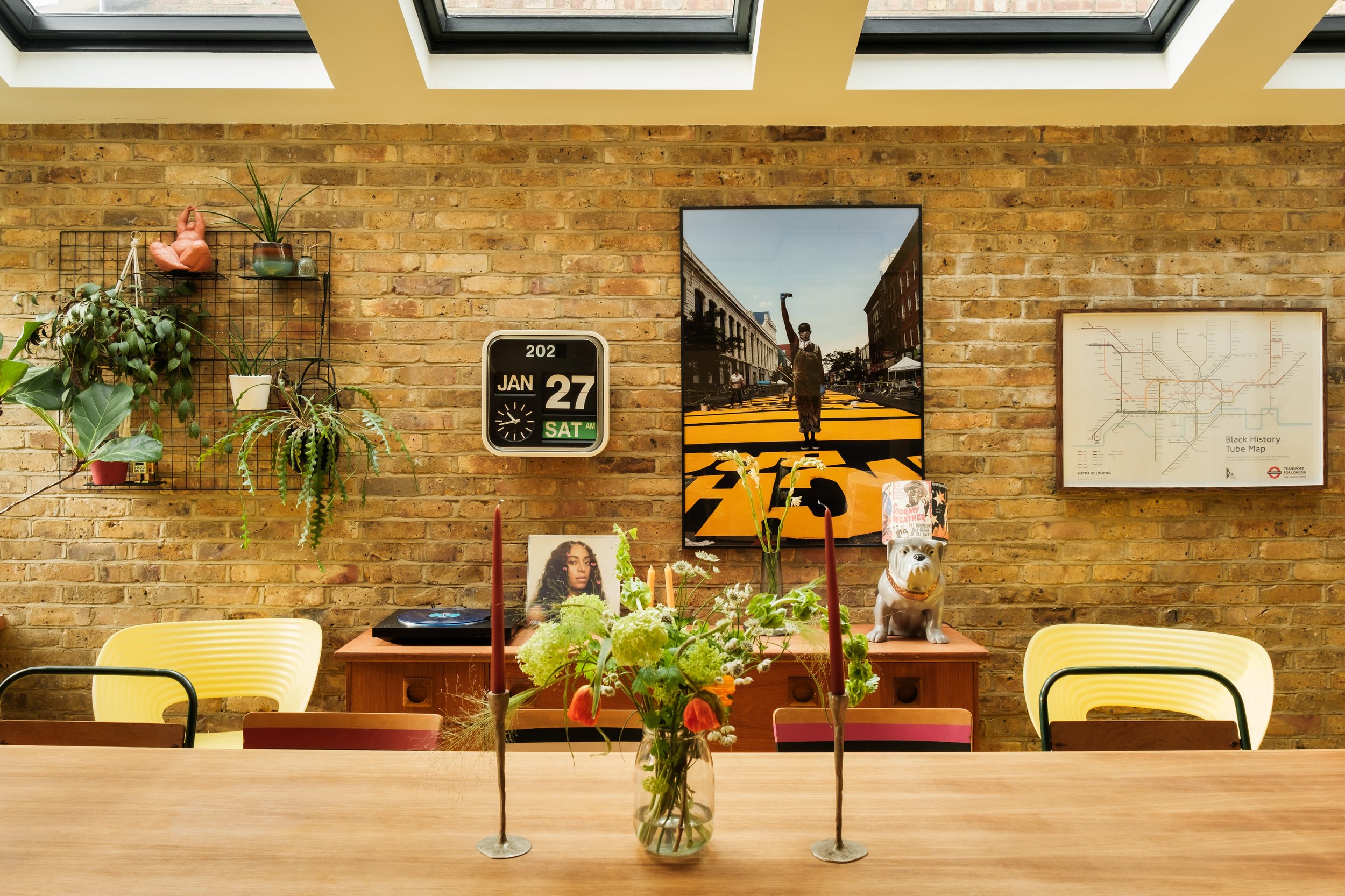 A cozy dining room with a wooden table, yellow chairs, and a brick wall decorated with artwork, plants, and a clock. The table has a flower arrangement and two red candles. There are skylights above letting in natural light.