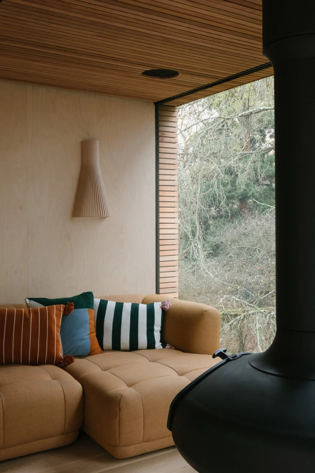 Living room corner with a beige sectional sofa, colorful striped and solid pillows, large window with wooden slats, a wall-mounted wooden light fixture, and a black stove in the foreground.