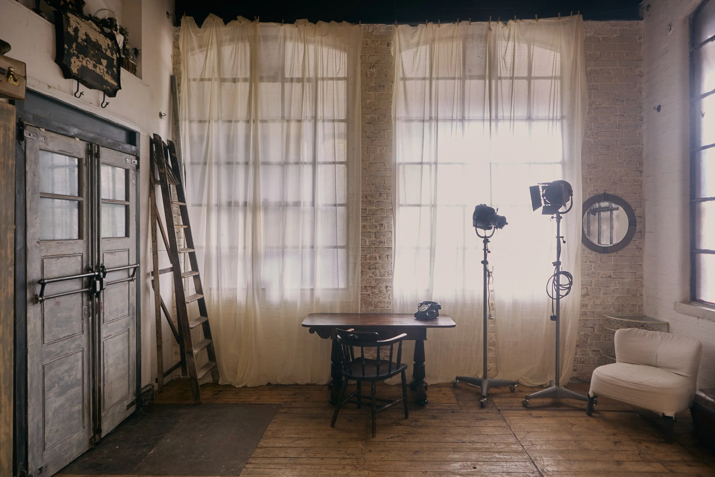 A studio or office space with large windows covered by sheer curtains, a wooden table with a vintage telephone, two studio lights on stands, an armchair, a round mirror, a ladder, and an old cabinet.