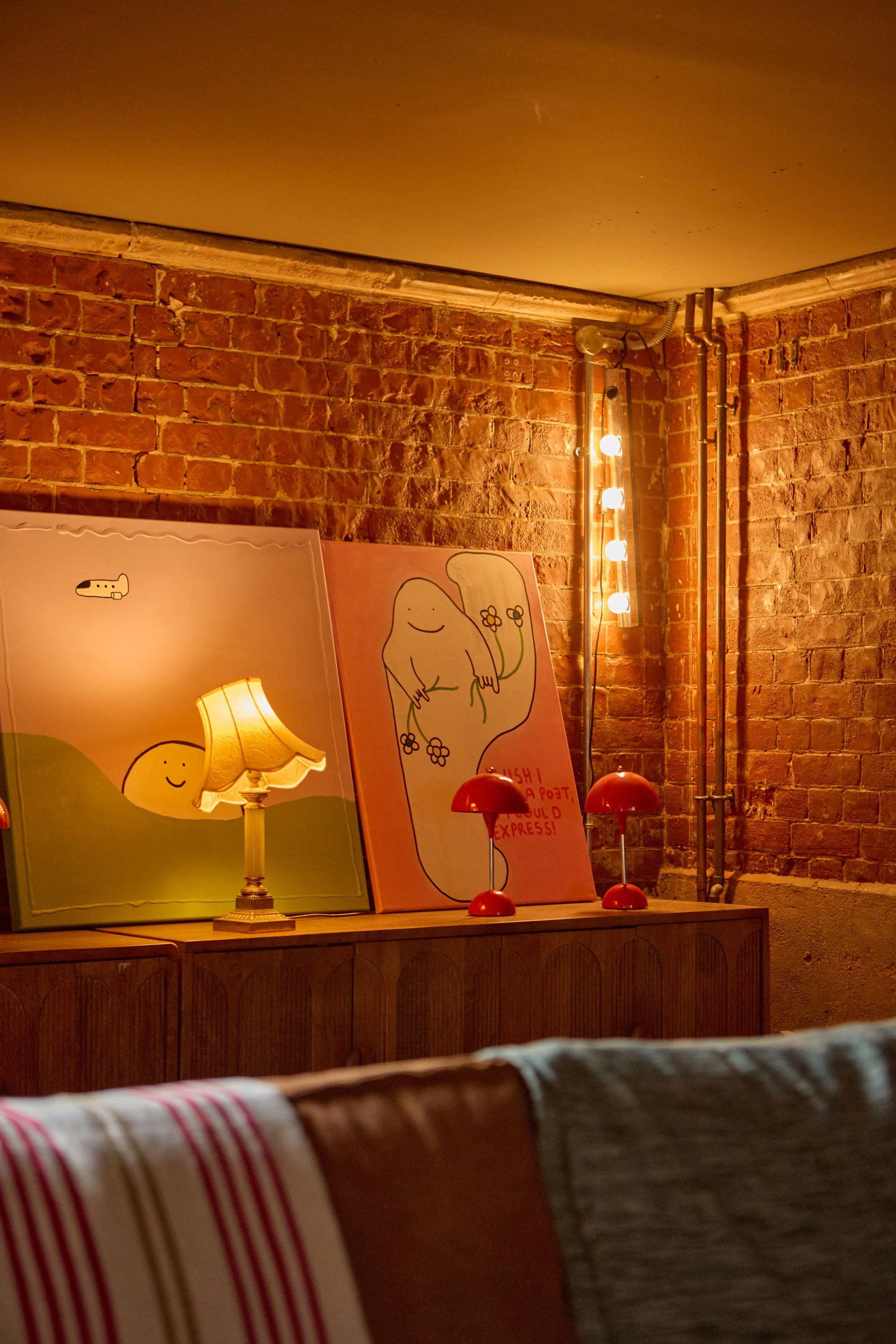 Interior scene with brick wall, two colorful paintings with simple cartoon figures, a lit table lamp, two red table lamps, and a string of round lights mounted on the brick wall.