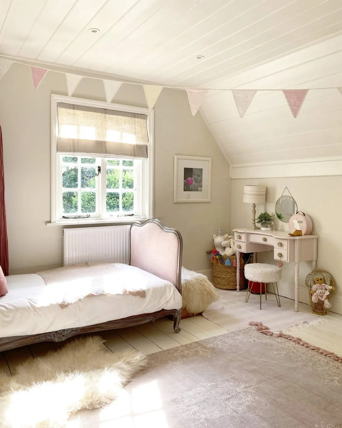 A cozy, feminine bedroom with a pink and white color scheme, featuring a bed with decorative pillows, a fluffy rug, a white dressing table with a mirror, and wall decorations including a framed picture and a bunting strip of pink and white fabric fla