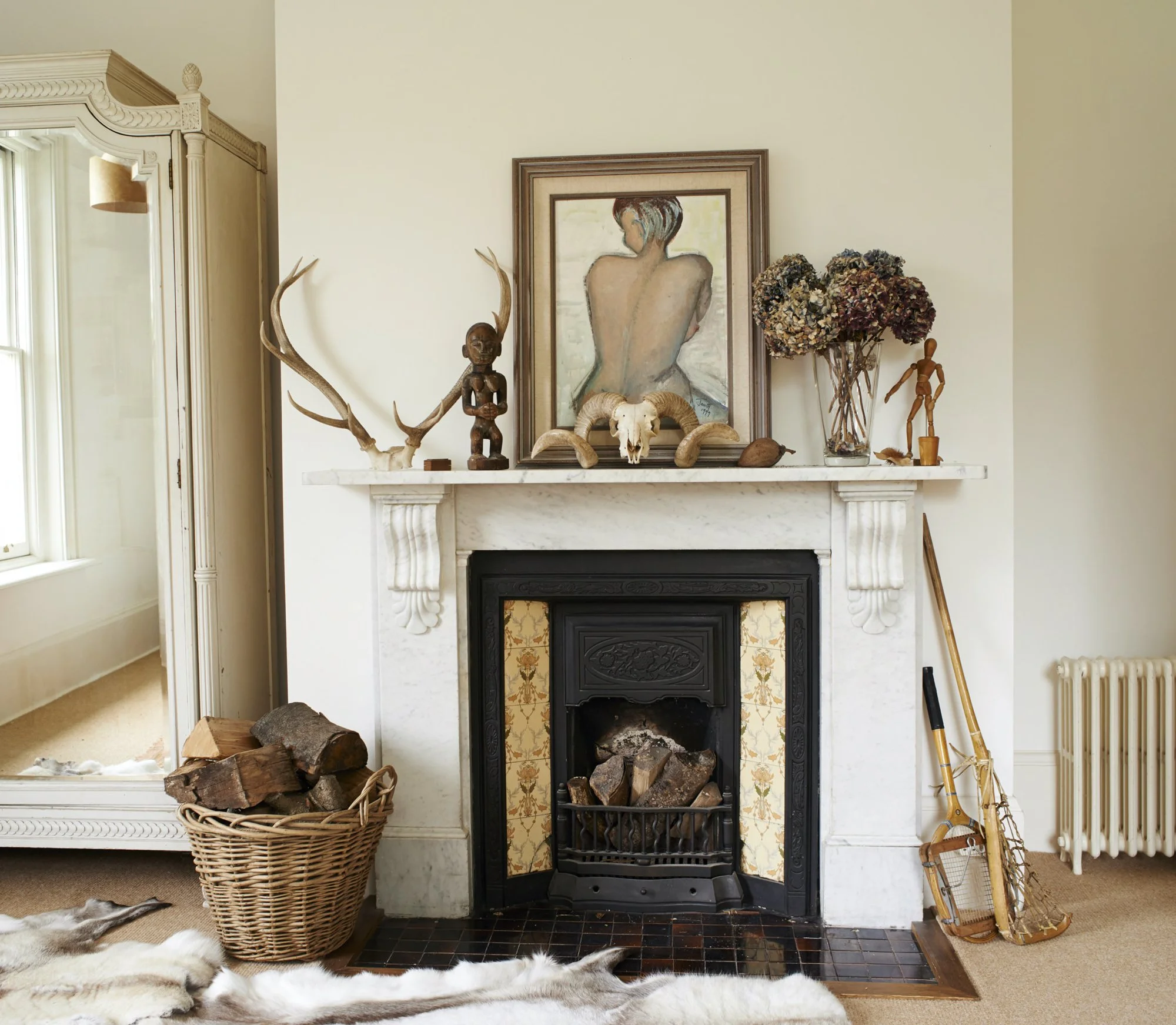 Living room fireplace with decorative items including antlers, a tribal doll, animal skull with horns, dried flowers, and a wooden sculpted figure. A portrait of a woman hangs above the mantel. A basket with logs, a mirror, and snowshoes are nearby.