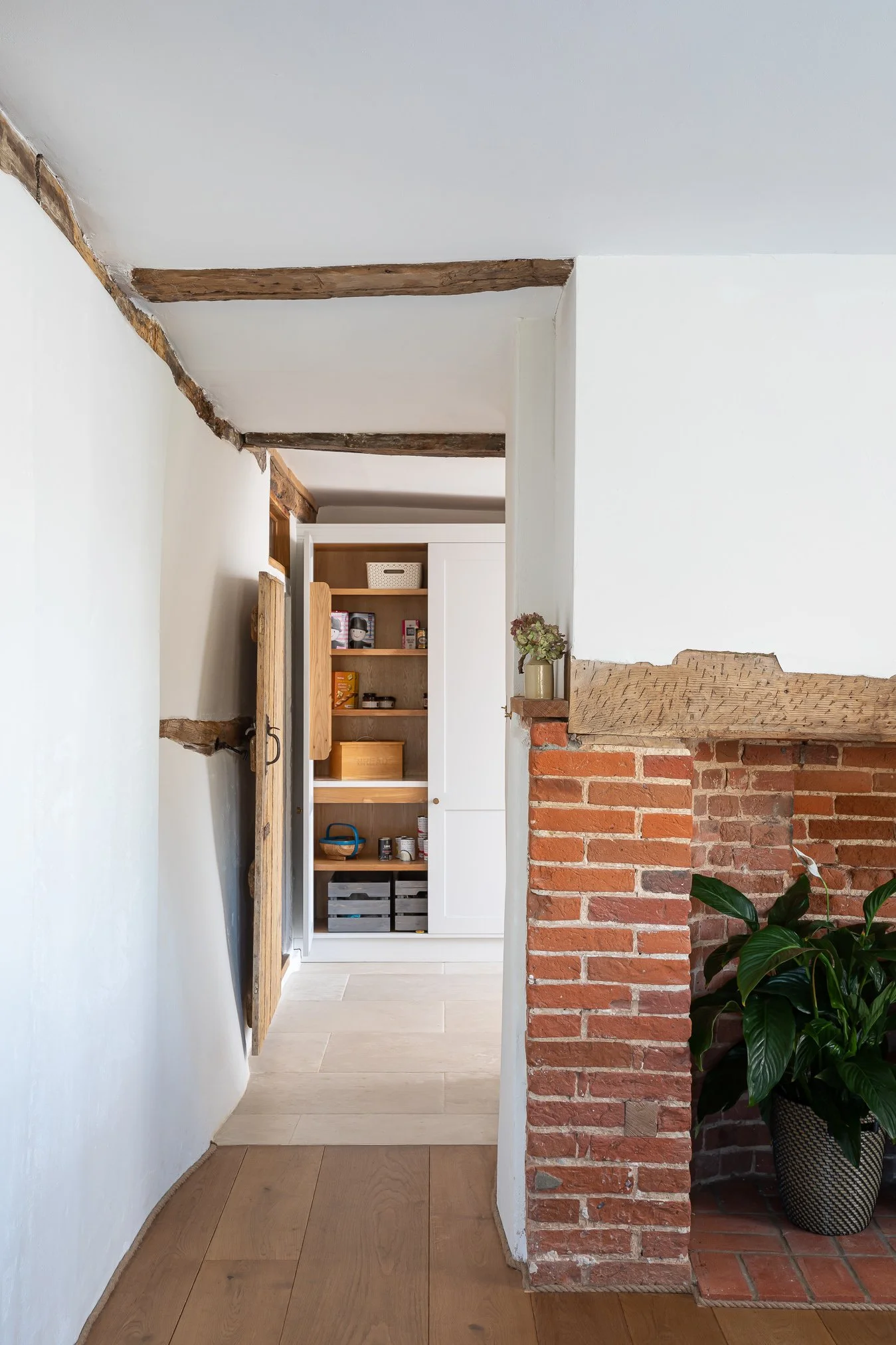 View of a cozy home interior featuring exposed wooden beams on the ceiling, a white storage cabinet with open shelves, a potted plant beside a brick fireplace, and warm wooden flooring.