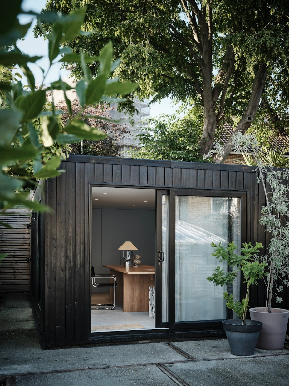 A small modern black wooden garden shed with a sliding glass door, interior includes a wooden desk, lamp, and chair, surrounded by potted plants and trees.