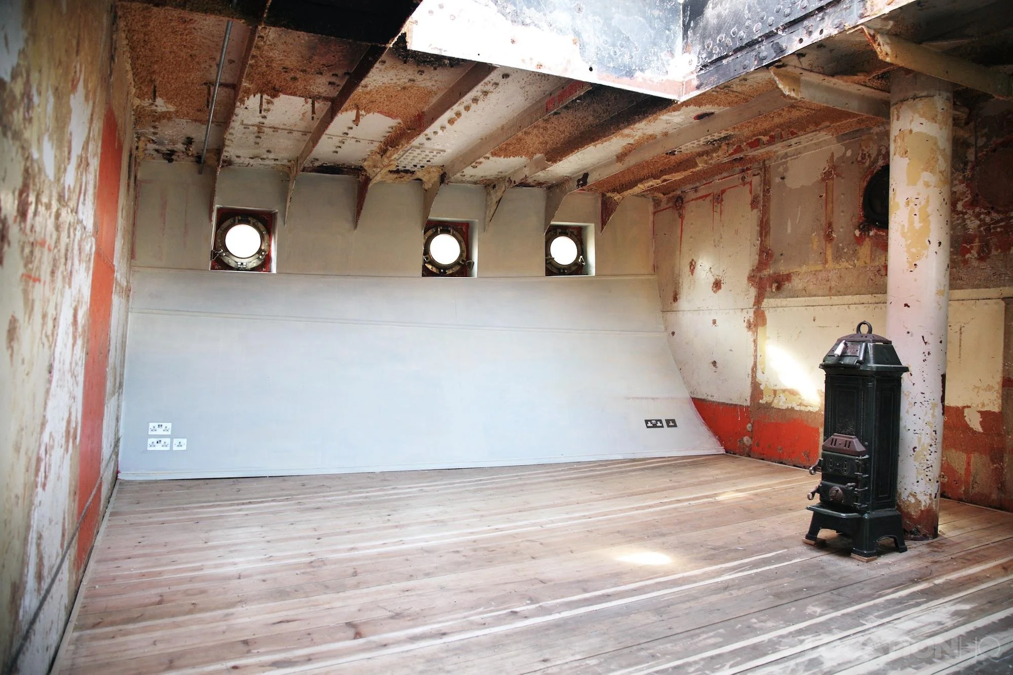 Unfinished room with exposed walls and ceiling, four round small windows, a wood stove in the corner, and electrical outlets on the wall.