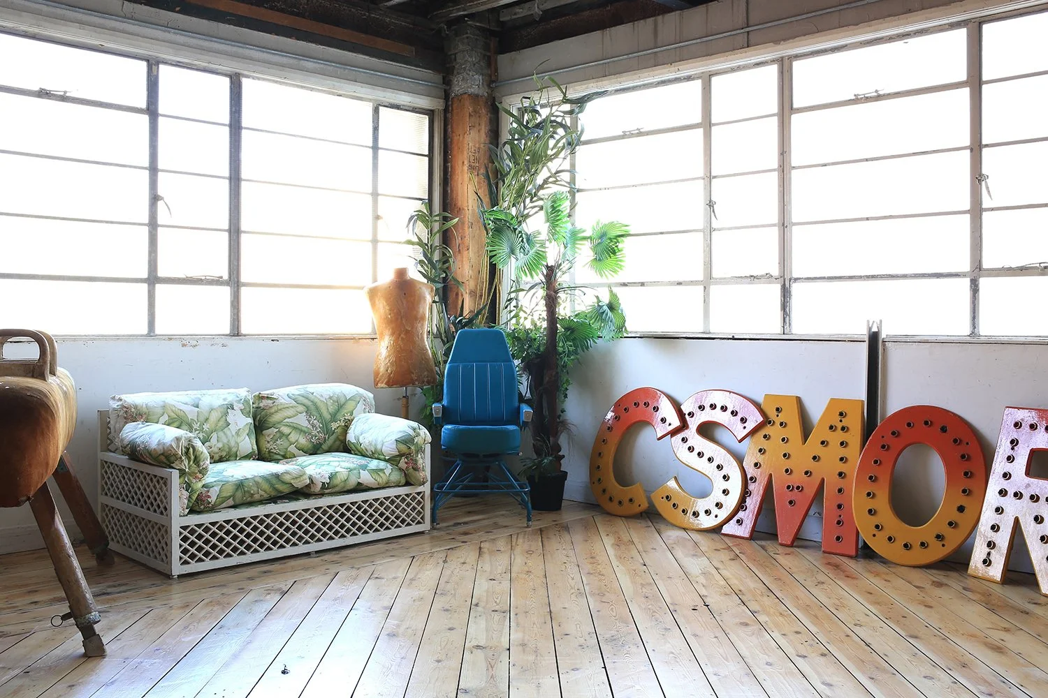 An interior room with large windows, a green leaf patterned sofa, a blue chair, potted plants, a dress form, and large vintage carnival-style letters spelling 'CASINO'.