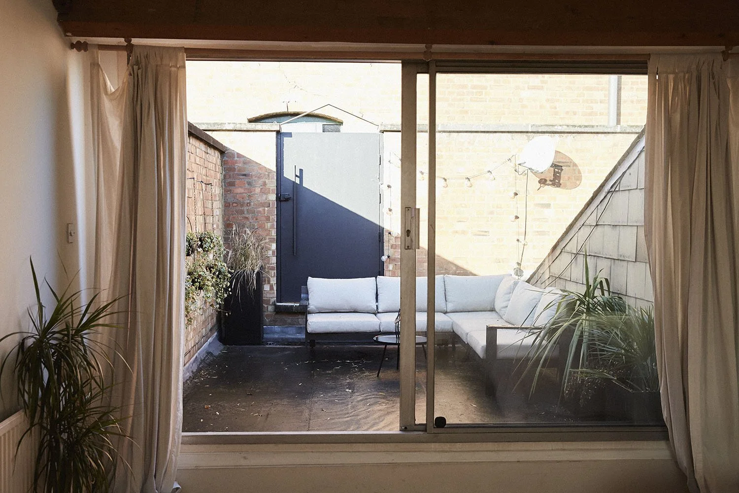 View of a cozy outdoor patio or balcony area with white cushioned seating, potted plants, and string lights, seen through large sliding glass doors with curtains.