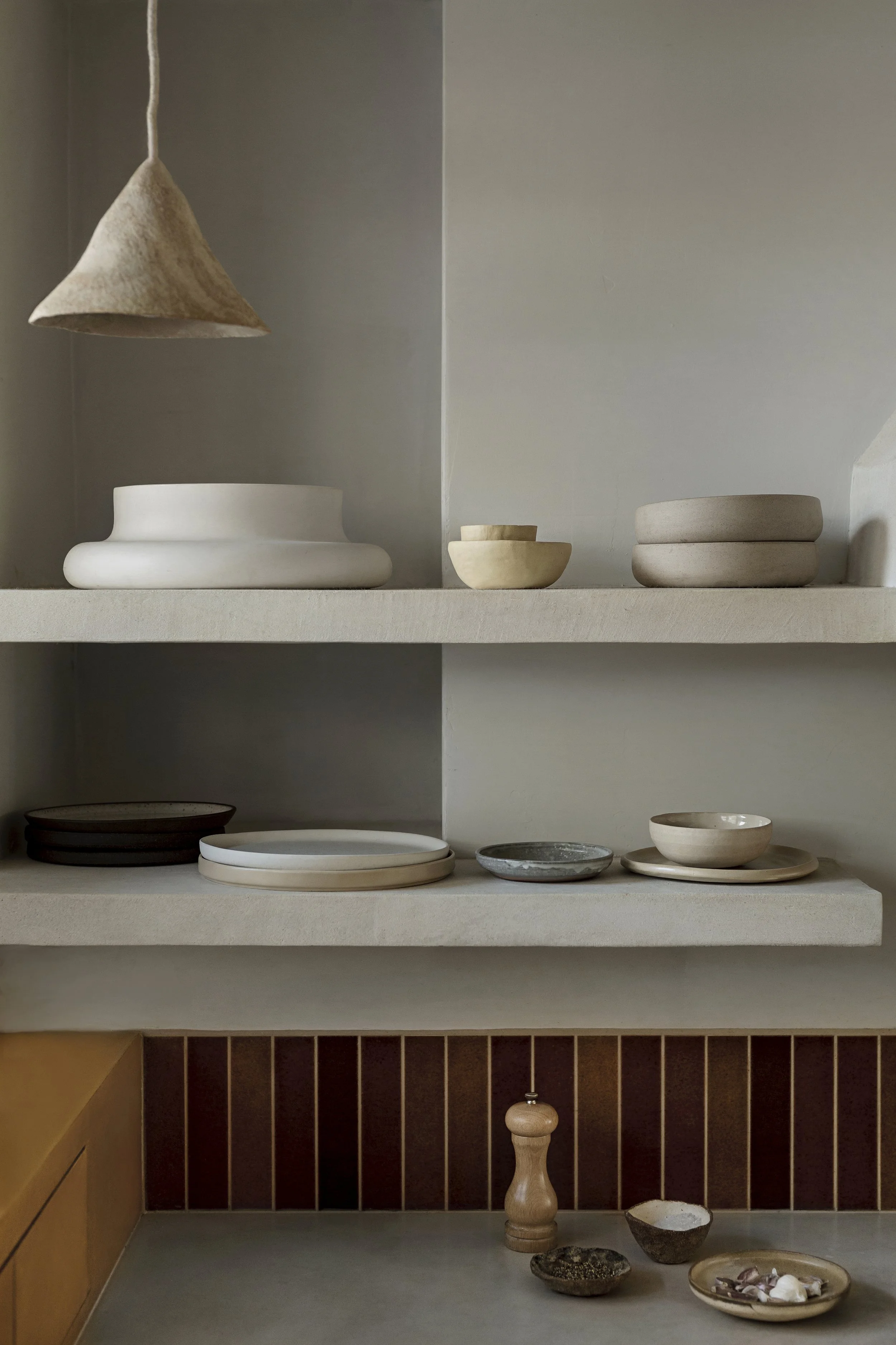 A neutral-colored display shelf with various ceramic dishes, bowls, and plates, with a small assortment of decorative stones and a pepper mill on the bottom shelf.