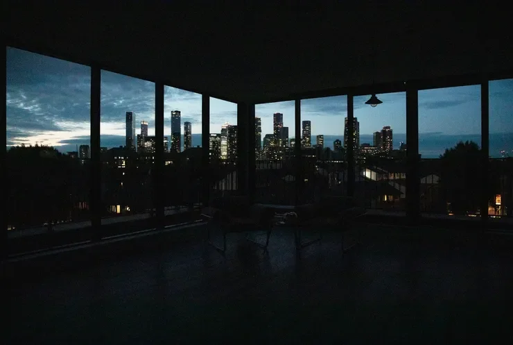 Nighttime view of a city skyline with tall buildings seen through large windows from inside a dark room or balcony.
