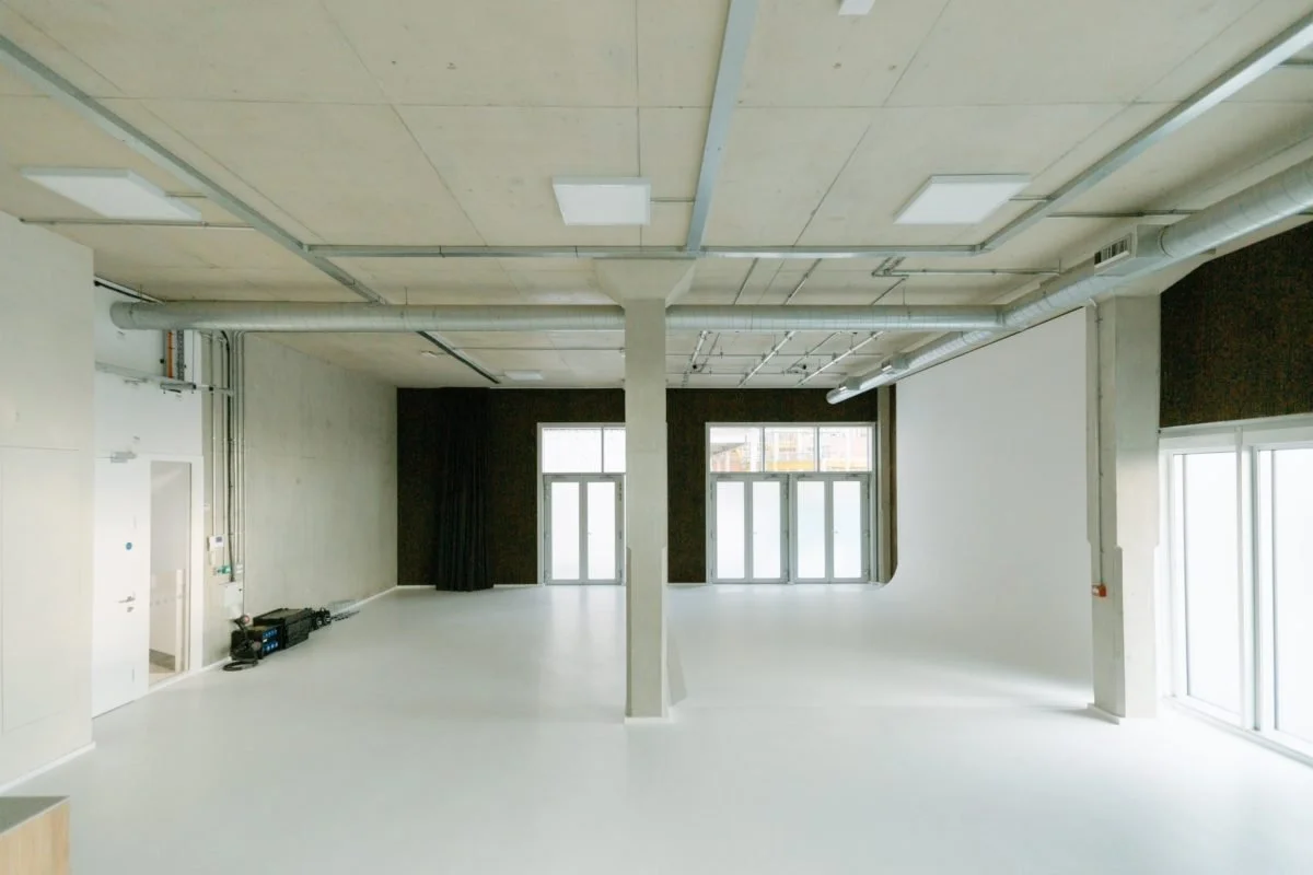Empty modern room with large windows, white walls, exposed ceiling pipes, and a light-colored floor.