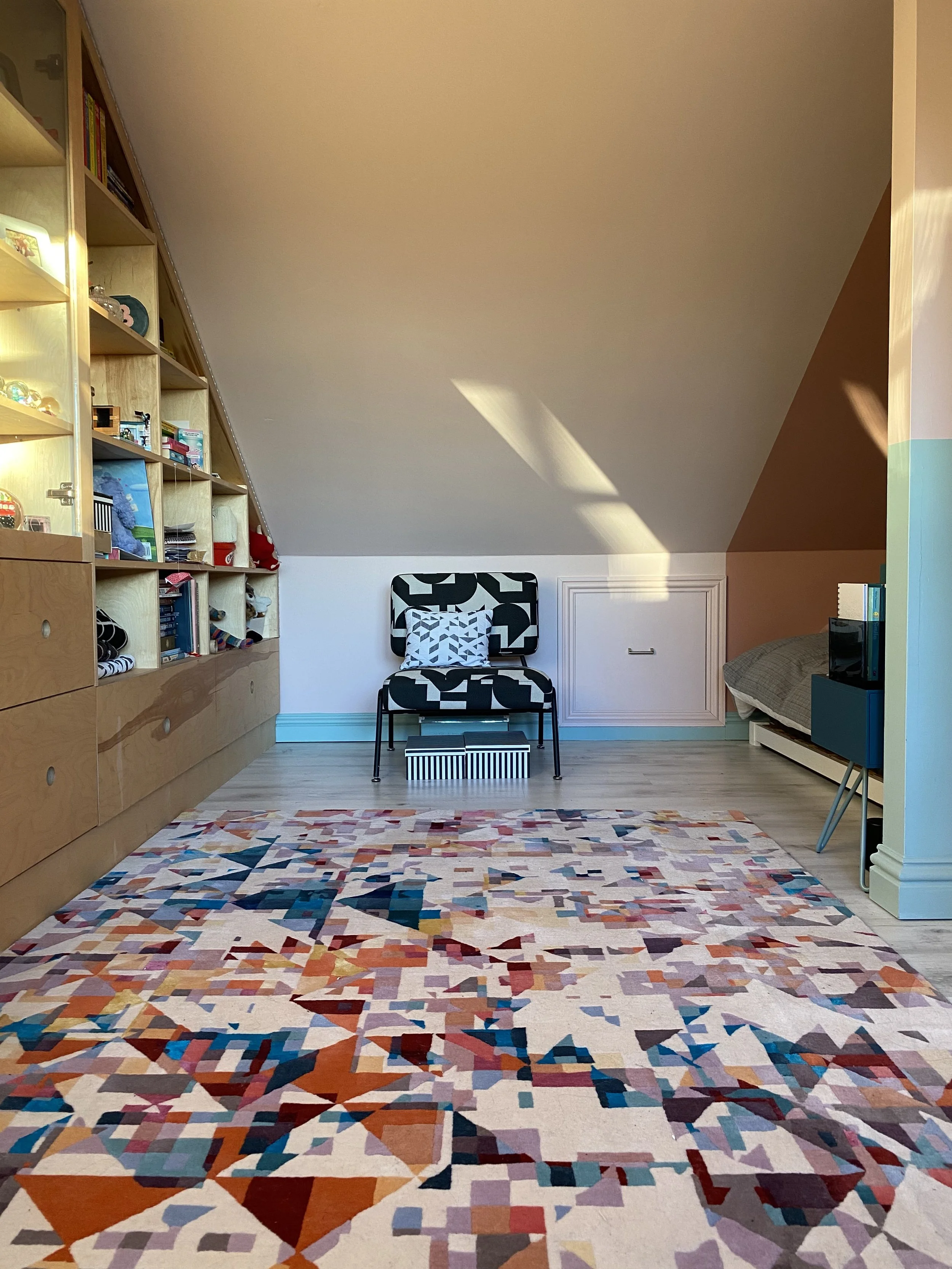 A cozy attic room with a colorful abstract geometric patterned area rug, a black-and-white patterned armchair with a pillow, a built-in brown wooden shelving unit filled with books and decorative items, a blue side table with books and a TV, and a sm