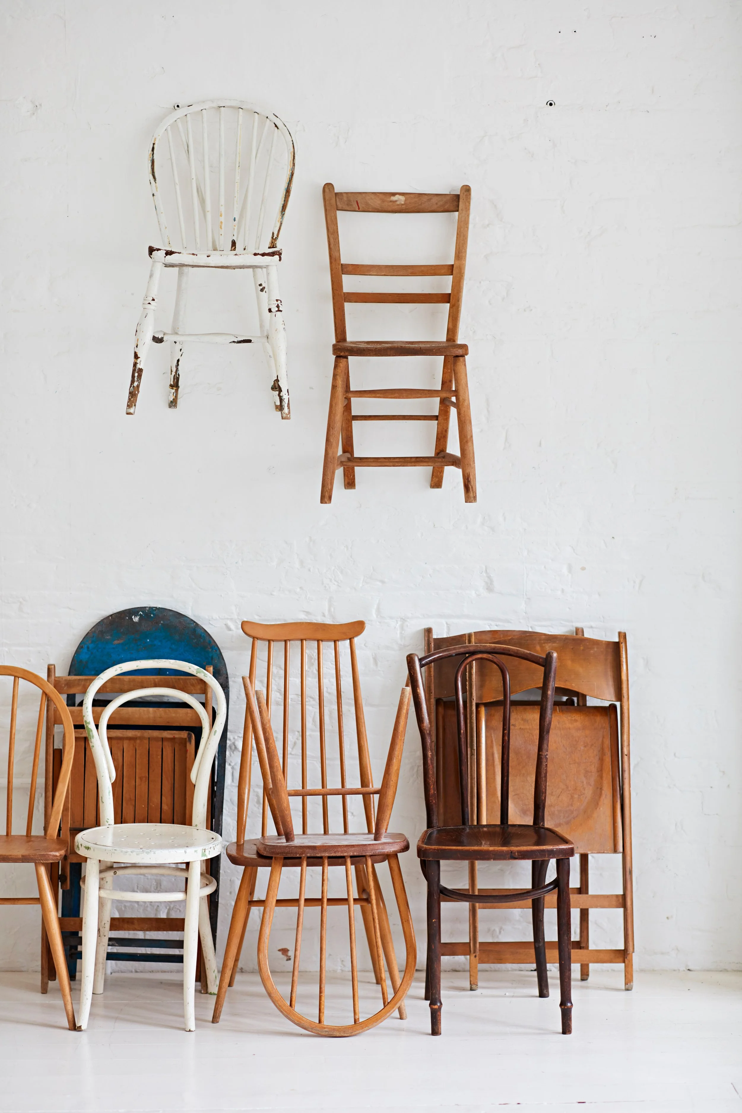 Various wooden and painted chairs and a bench arranged against a white brick wall, with some chairs stacked and others leaning.