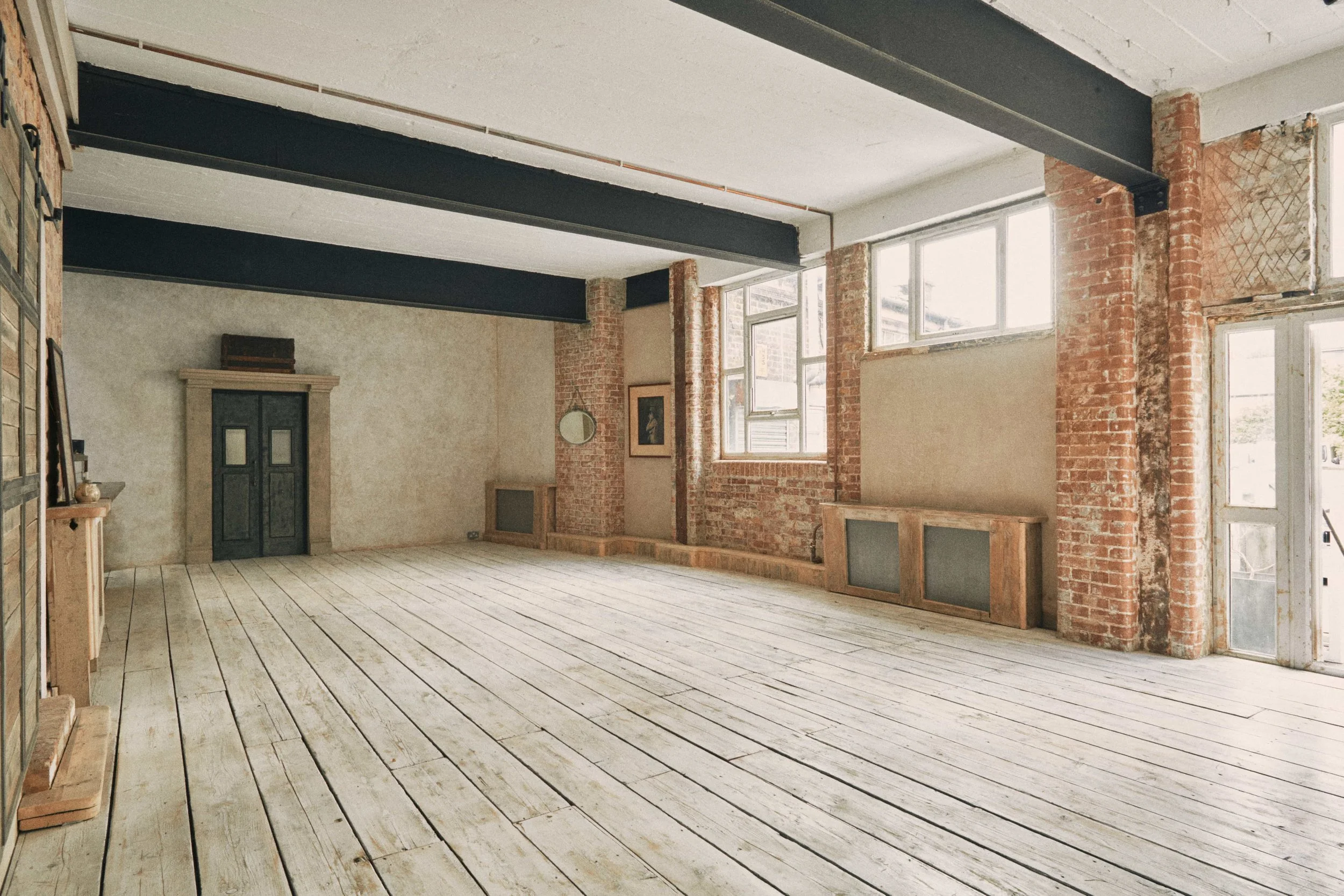 Empty industrial-style living room with exposed brick walls, white wooden floorboards, large windows, and a high ceiling with dark beams.