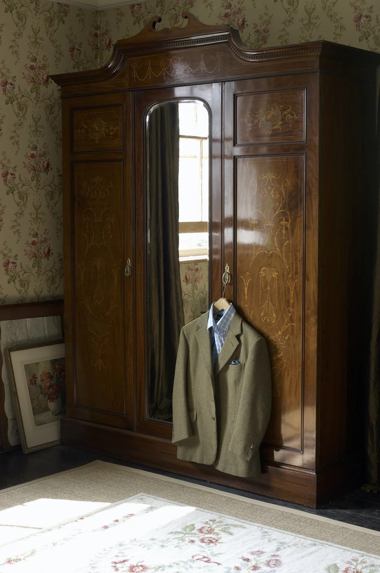An antique wooden wardrobe with floral inlay details, a mirror on one door, and a suit jacket hanging on the handle. The wardrobe is in a room with floral wallpaper, framed pictures resting on the floor, and a cream-colored rug with floral patterns, 