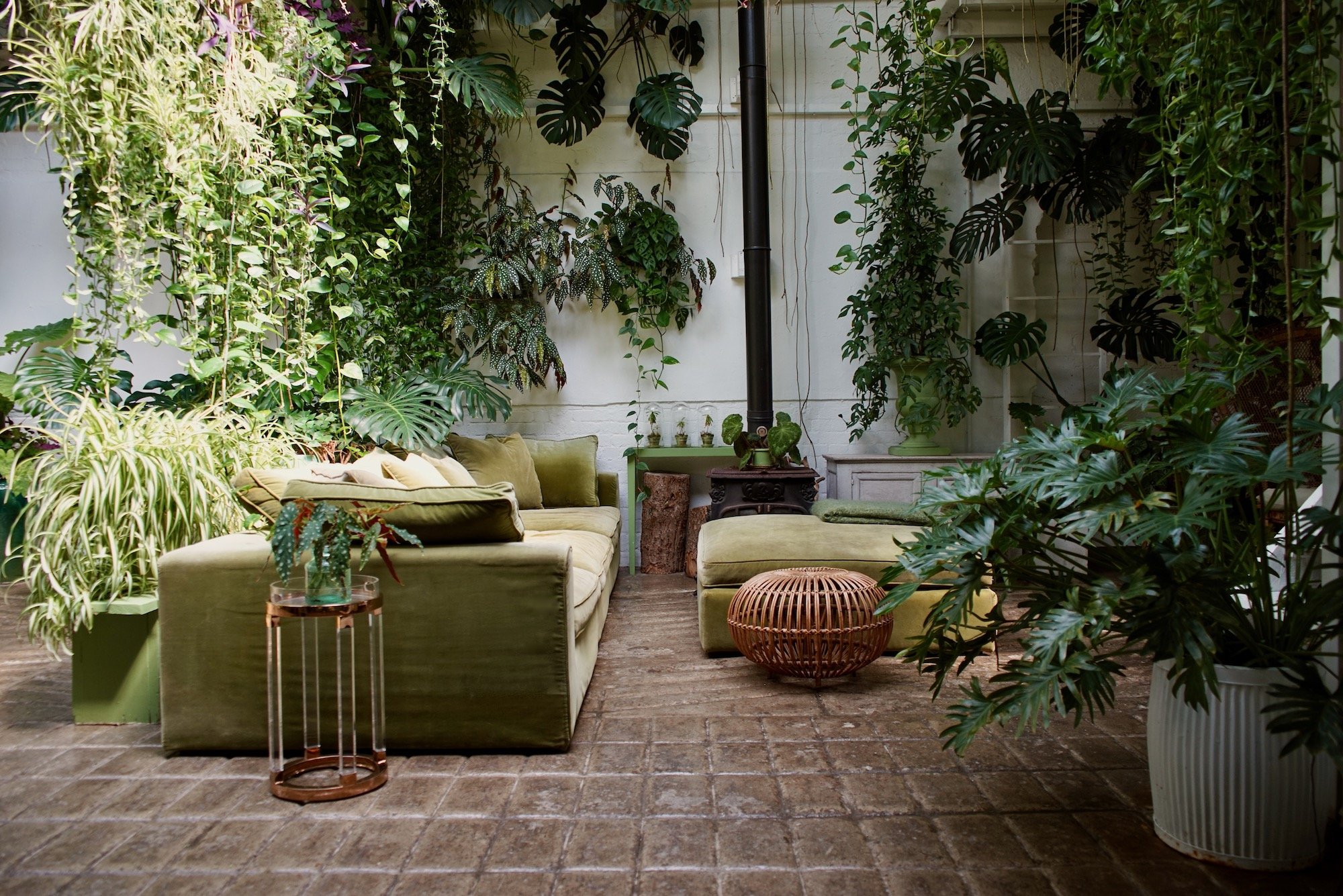 A cozy indoor garden with lush green plants and a vintage-style green sofa, a wicker ottoman, and a small round side table with a glass top and plant. The space features a brick floor and white walls.