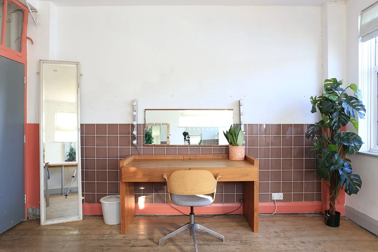 A minimalistic workspace with a wooden desk, a gray and wooden swivel chair, a wall mirror with light bulbs, a potted plant on the desk, a tall indoor plant in a pot to the right, a large window letting in natural light, and a full-length mirror lean
