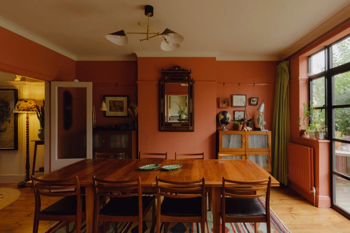 Dining room with a wooden table, black chairs, a sideboard, framed artwork, potted plants, and a large window with green curtains.