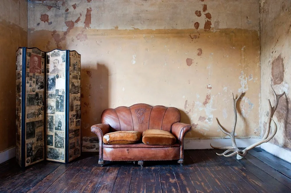 An old, brown leather sofa with two cushions on a wooden floor in front of a distressed, yellowed wall. To the left, there is a vintage photo collage folding screen. To the right, there are large, decorative antlers mounted on the floor.