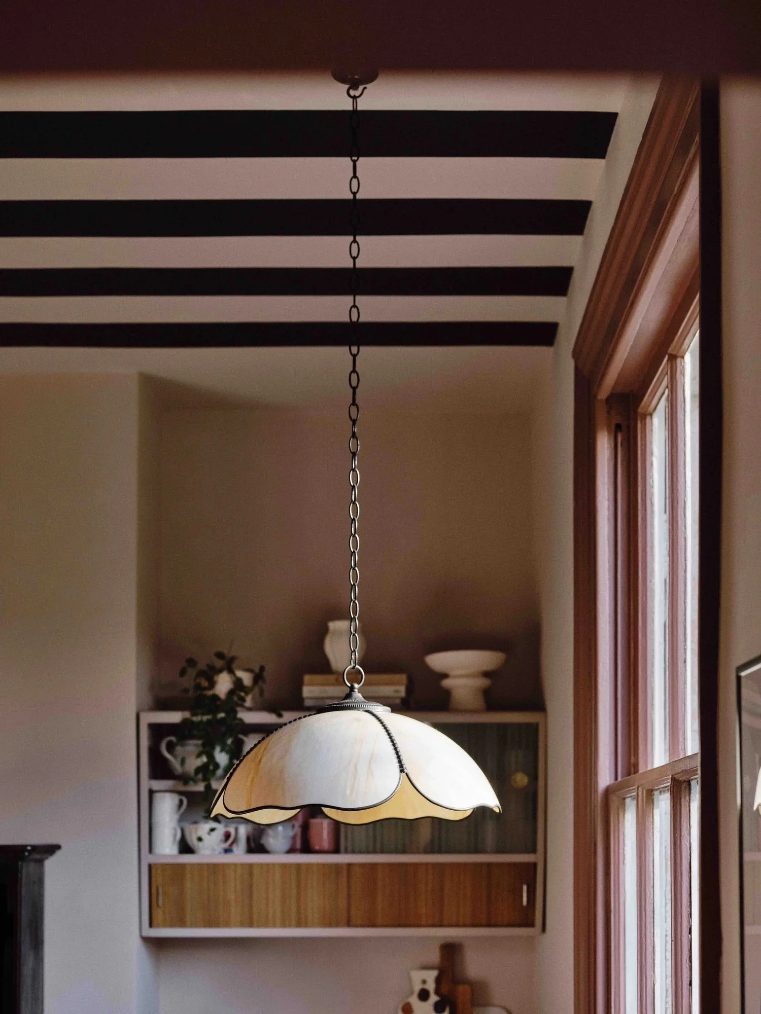 A hanging kitchen or dining area lamp with a scalloped, cream-colored glass shade and metal chain, positioned in front of a window with wooden trim and a wall cabinet with decorative items inside.