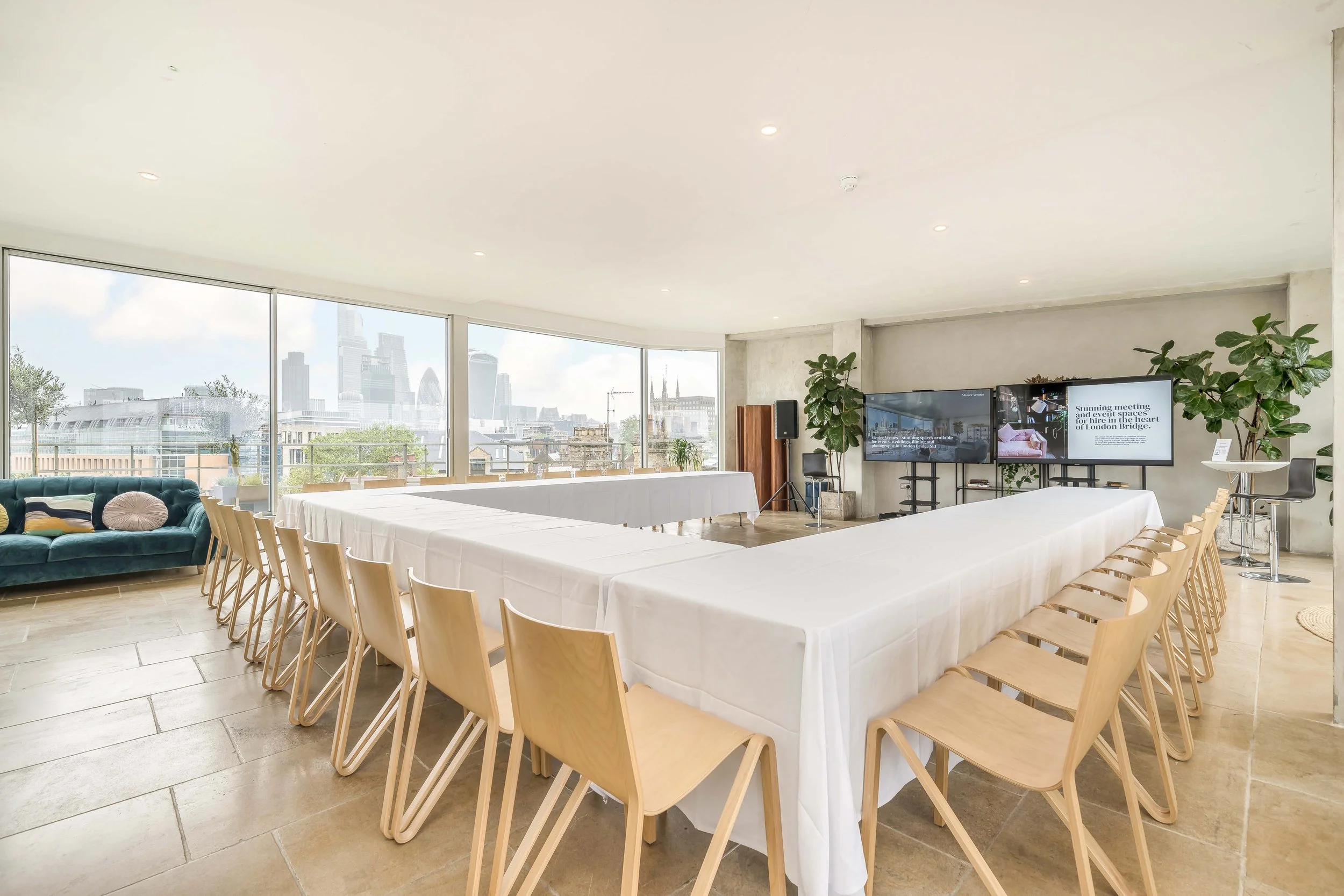 Conference room with U-shaped table, beige chairs, large windows with city skyline, TV screens, plants, and white walls.