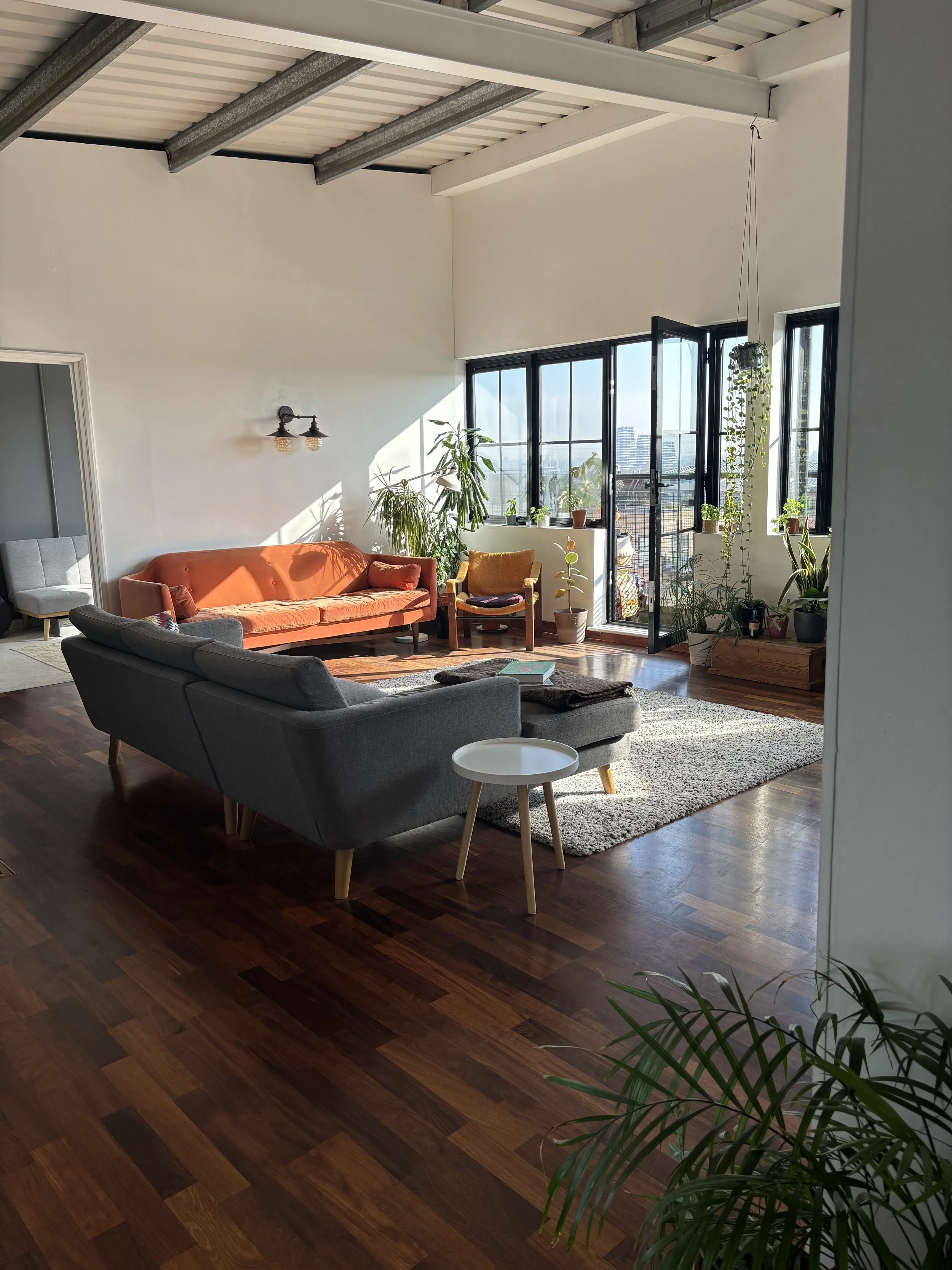 Living room with wooden floors, gray and orange couches, a cream rug, and large windows with potted plants, sunlight, and city view.