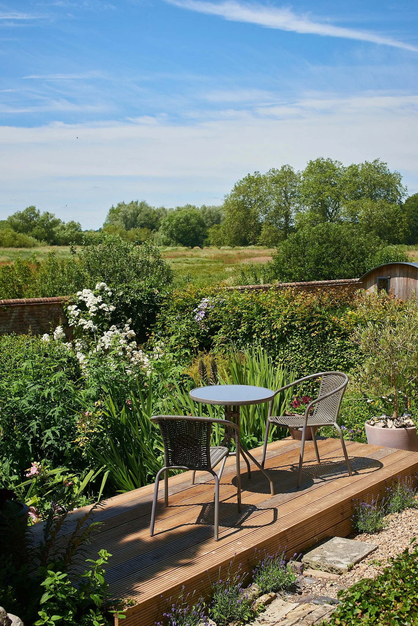 A cozy garden patio with a small metal table and two chairs on a wooden deck, surrounded by lush green plants and flowering bushes, overlooking a vast open field and a blue sky with wispy clouds.