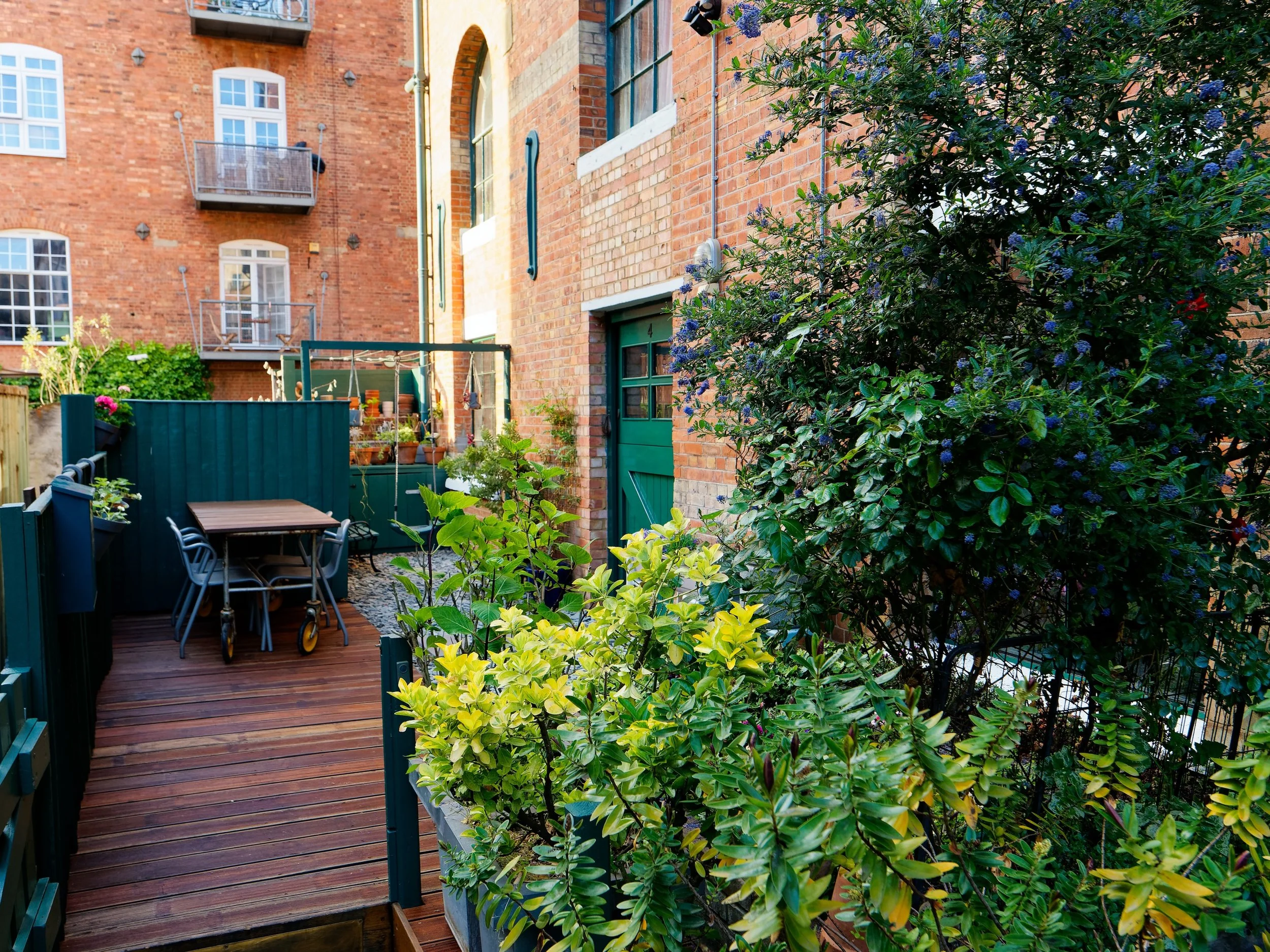 A cozy urban backyard patio with wooden decking, potted plants, a dinner table with chairs, and lush greenery surrounded by brick buildings.