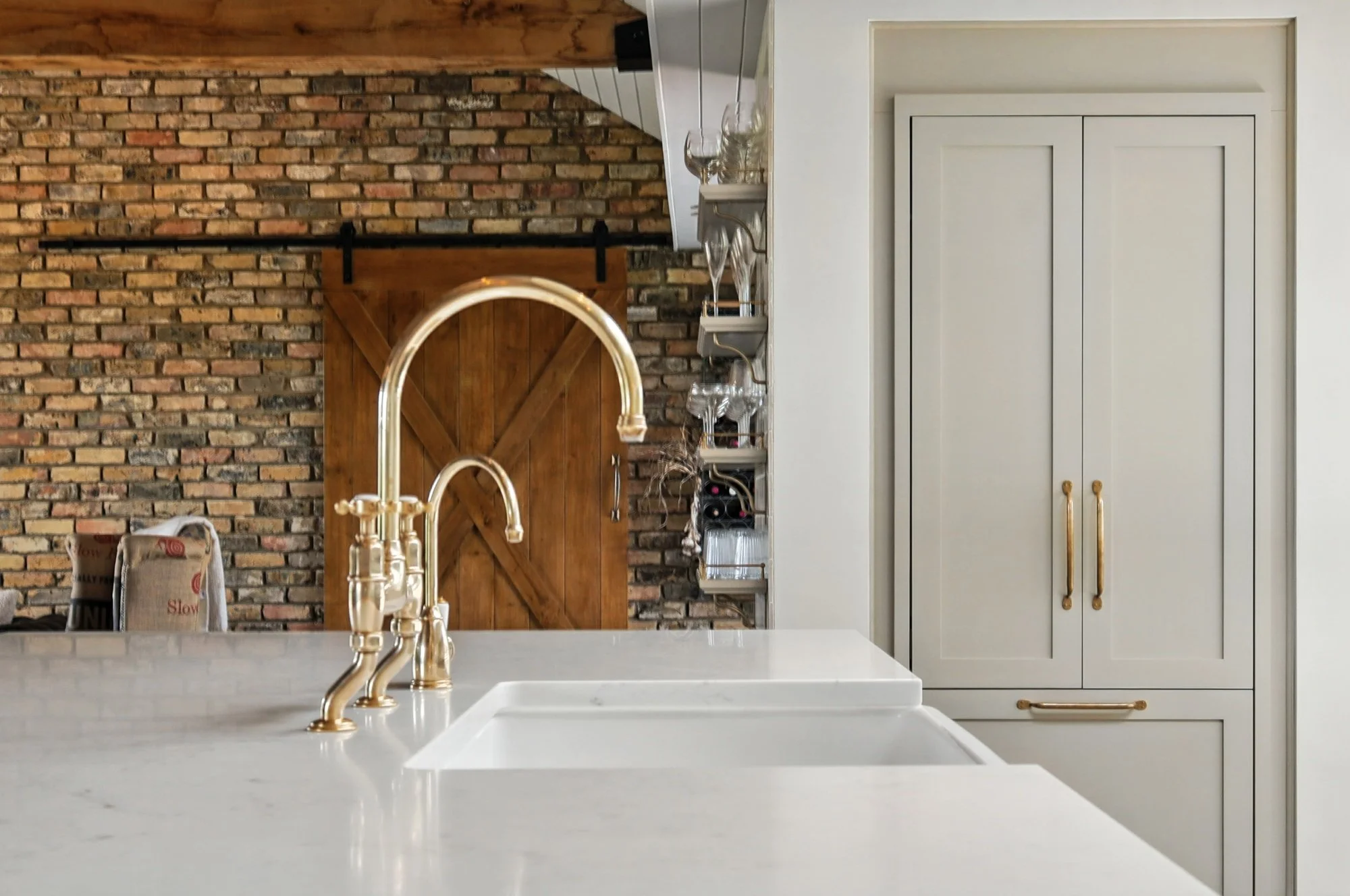 Modern kitchen with a white marble countertop, gold sink faucet, white cabinets with gold handles, a wine rack, and a brick wall with a sliding barn door.