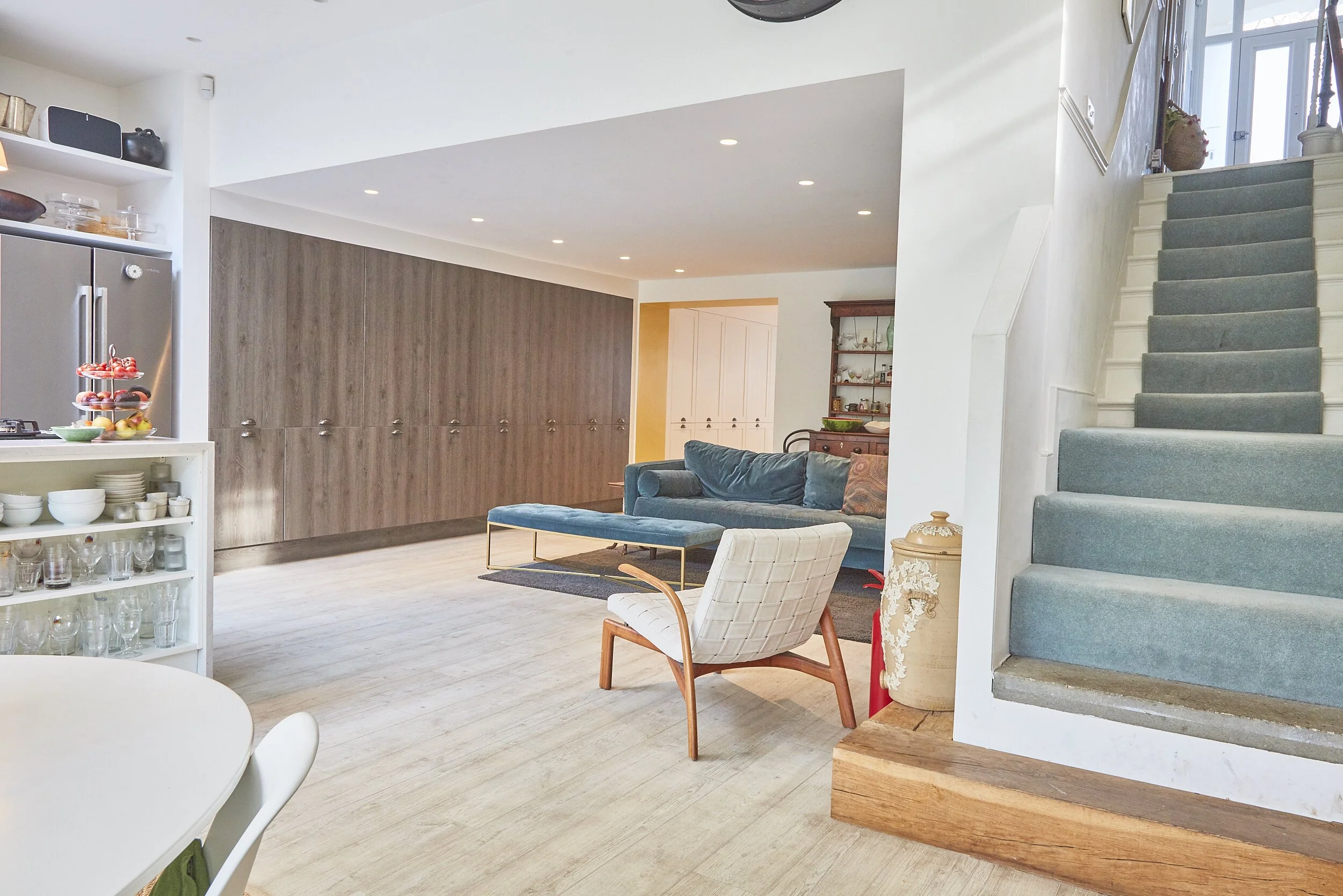 Living room with modern furniture, including a blue sofa, white and wooden armchair, and a staircase with green carpeted steps.
