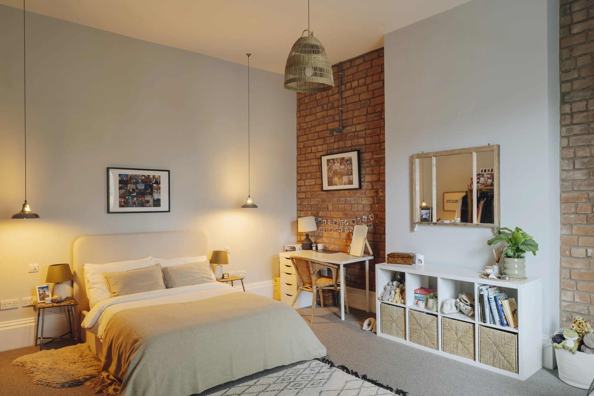 A cozy bedroom with a bed, side tables with lamps, a desk with a chair, a white shelving unit, and exposed brick wall accents, decorated with framed photos, a mirror, and plants.