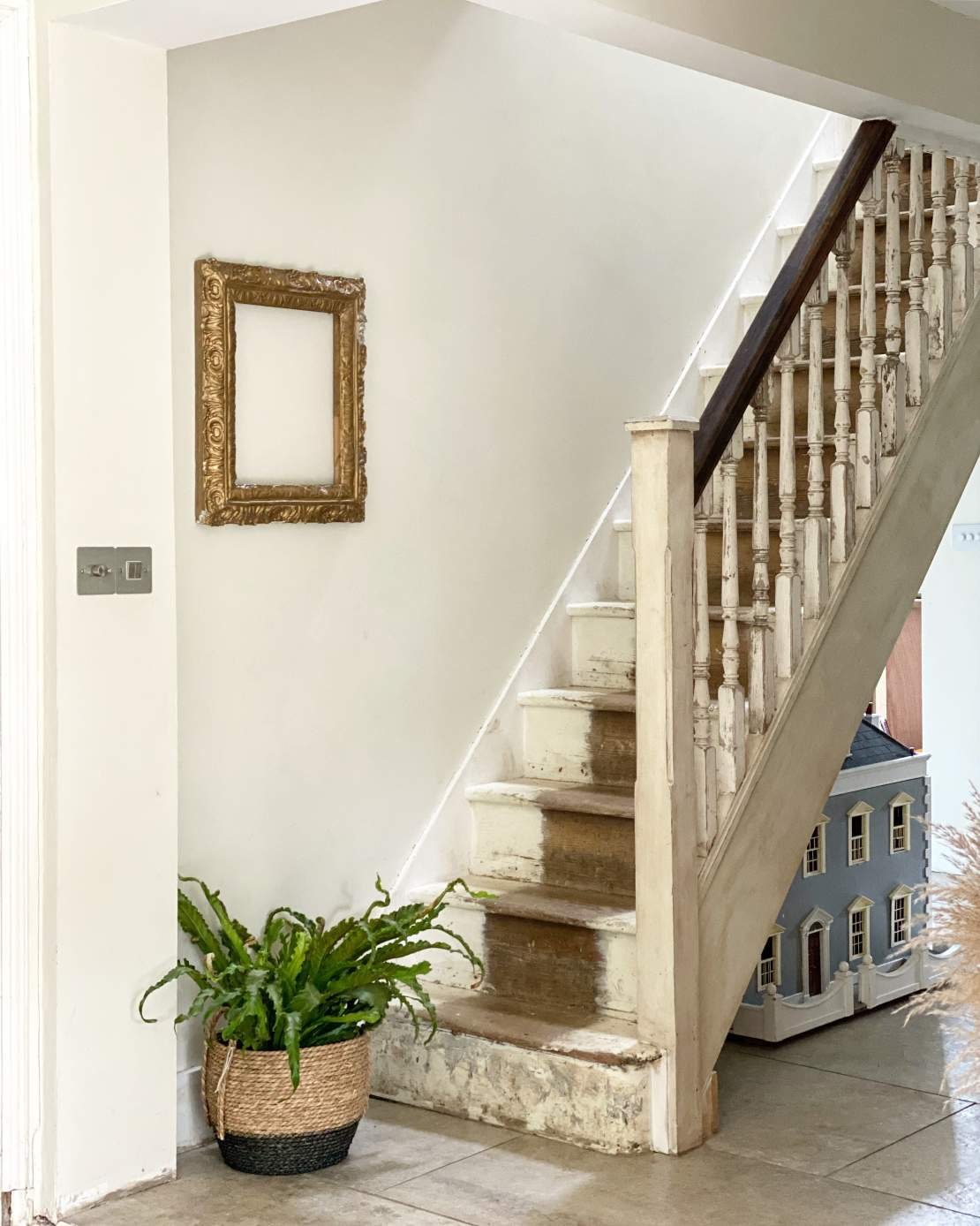 A staircase with worn, white-painted wooden steps and a dark handrail, next to a wall with a gold empty picture frame and a potted green plant in a woven basket.