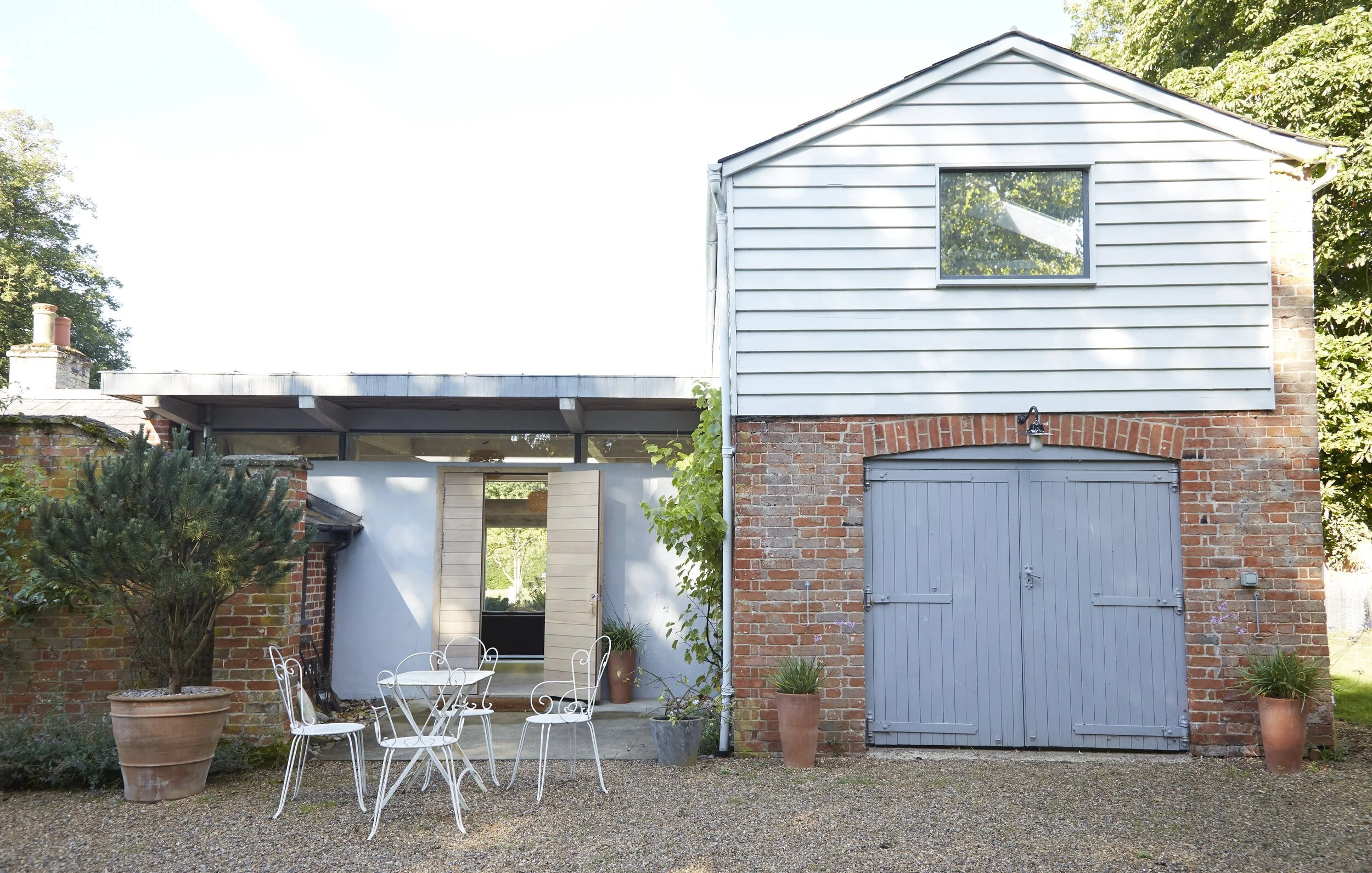 Exterior view of a brick and wood house with a yard. The house has a large garage door, a window on the upper level, and a side door. There are potted plants and a white metal outdoor dining set with chairs in the yard.