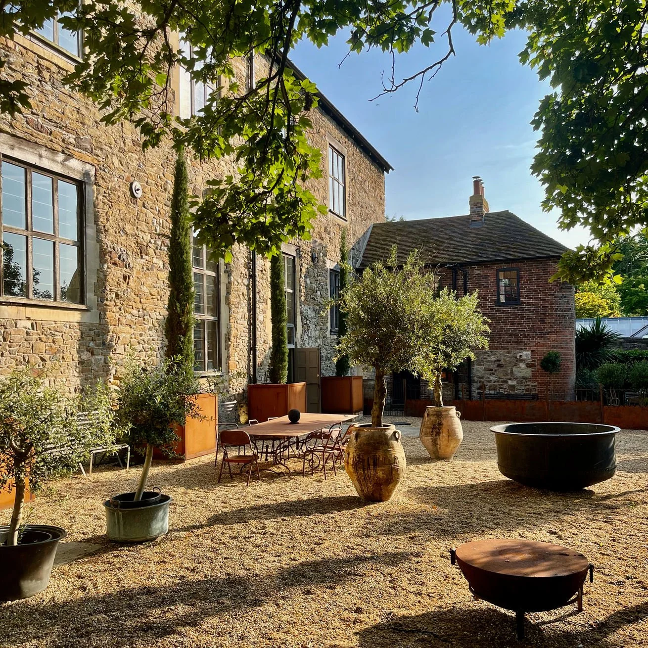 A courtyard with large potted trees, a table with chairs, and old brick buildings, under a blue sky and shade from a tree.