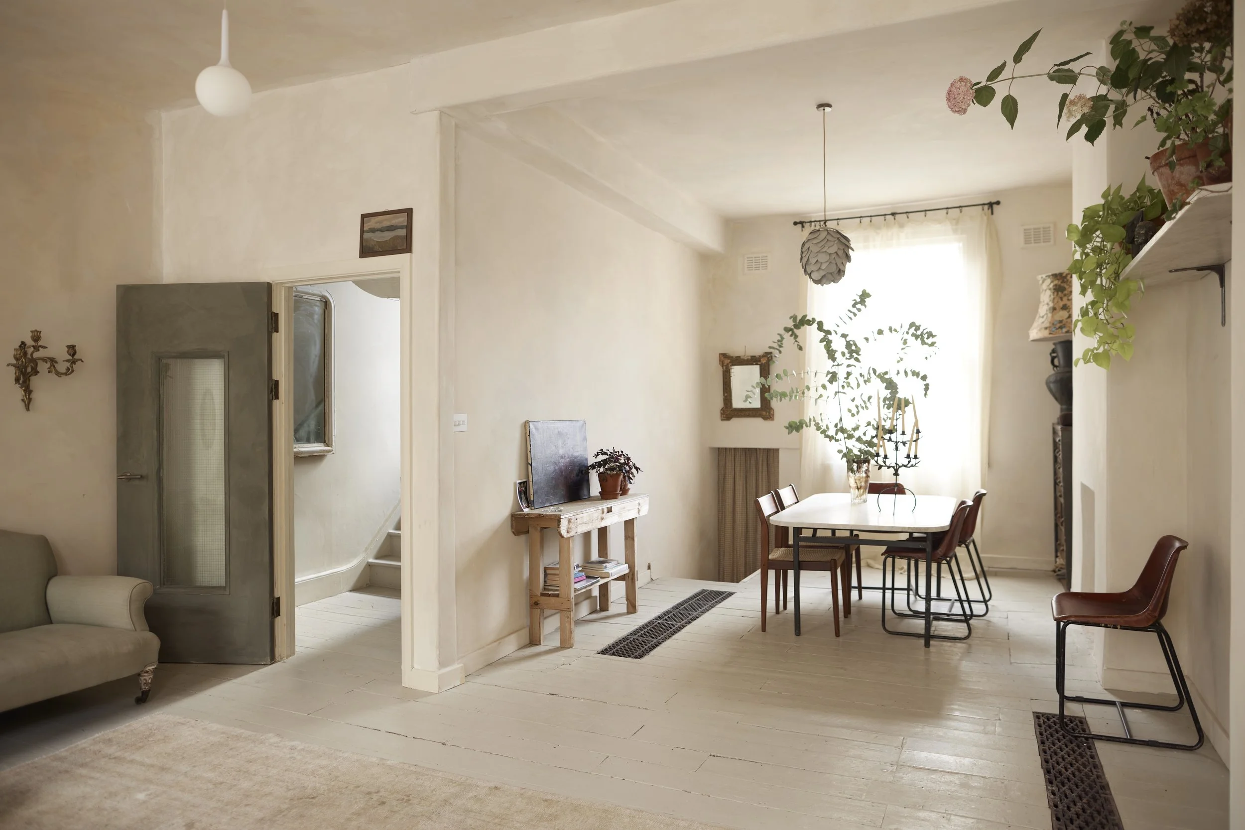 Living and dining area with beige walls, white wooden floor, a sofa, a small TV on a rustic stand, a dining table with chairs, potted plants, and decorative items like a mirror and artwork.