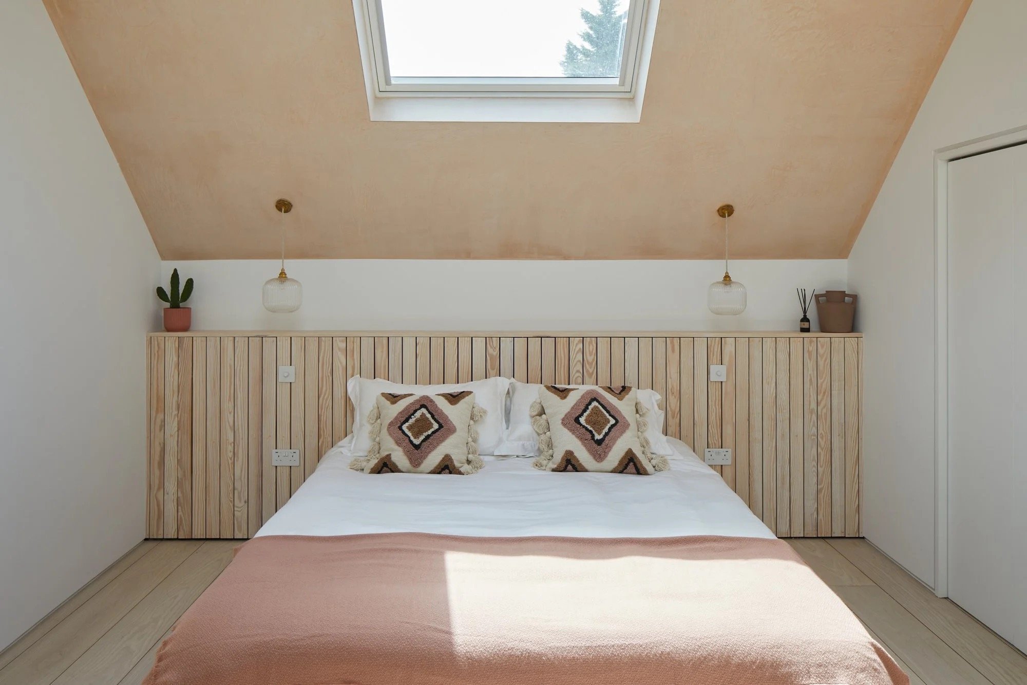 A cozy bedroom with a large bed, white bedding, and decorative pillows featuring geometric patterns. The room has a wooden headboard, a sloped ceiling with a skylight, and minimalist decor including a potted cactus, a small package, and reed diffuser