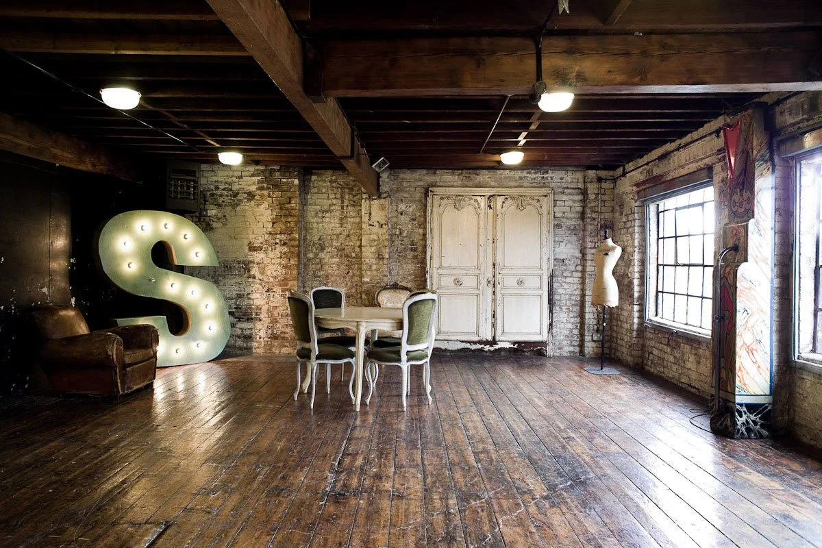 An industrial-style loft with exposed brick walls, wooden beams, and hardwood floors. There is a large light-up dollar sign, a vintage armchair, a white dining table with four chairs, a dress form, and a motorcycle in the corner near big windows.