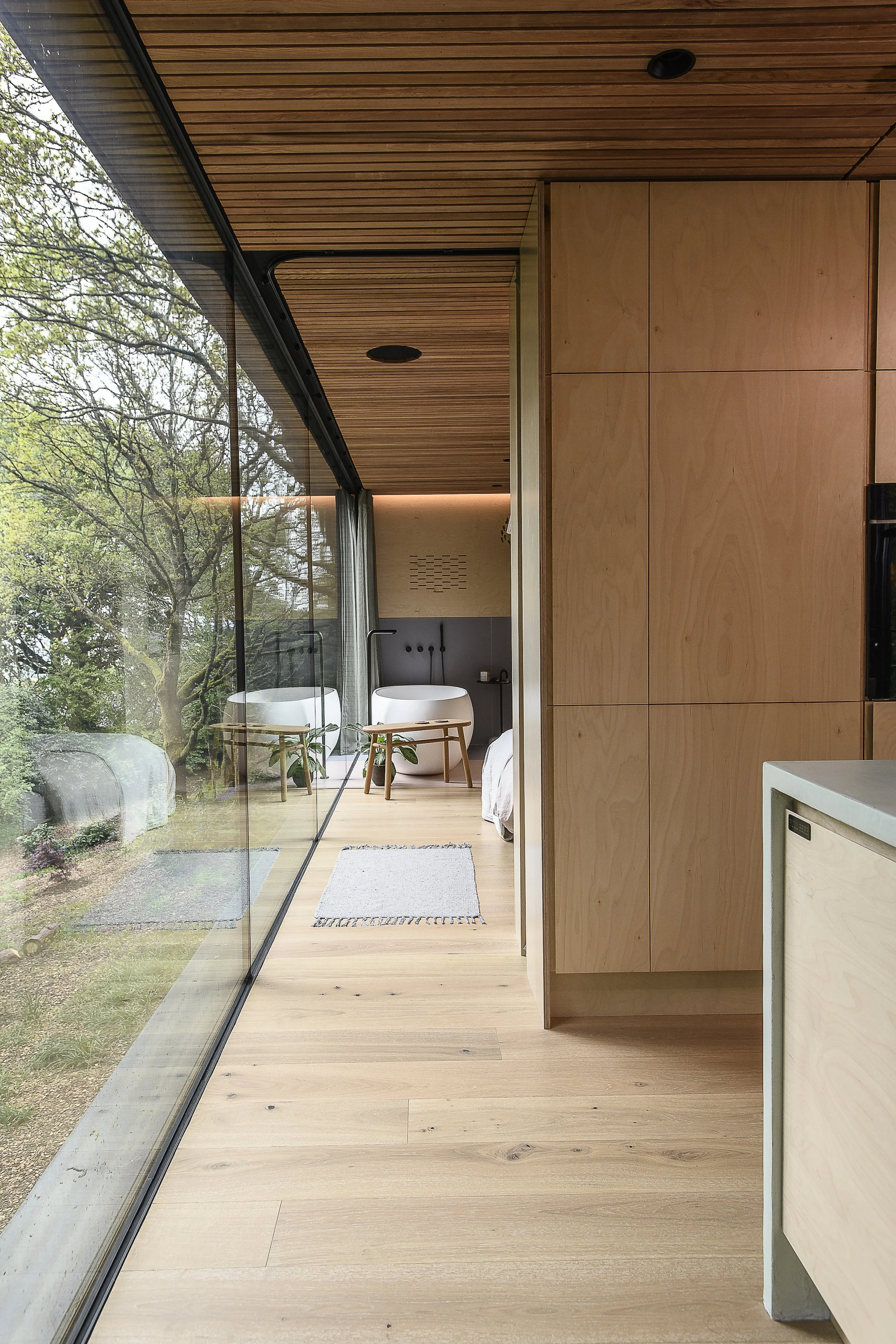A modern interior room with wooden flooring, light wood cabinetry, and a large glass wall showing a view of green trees outside. There is a white bathtub with a small wooden stool in front of it, and a bed partially visible.