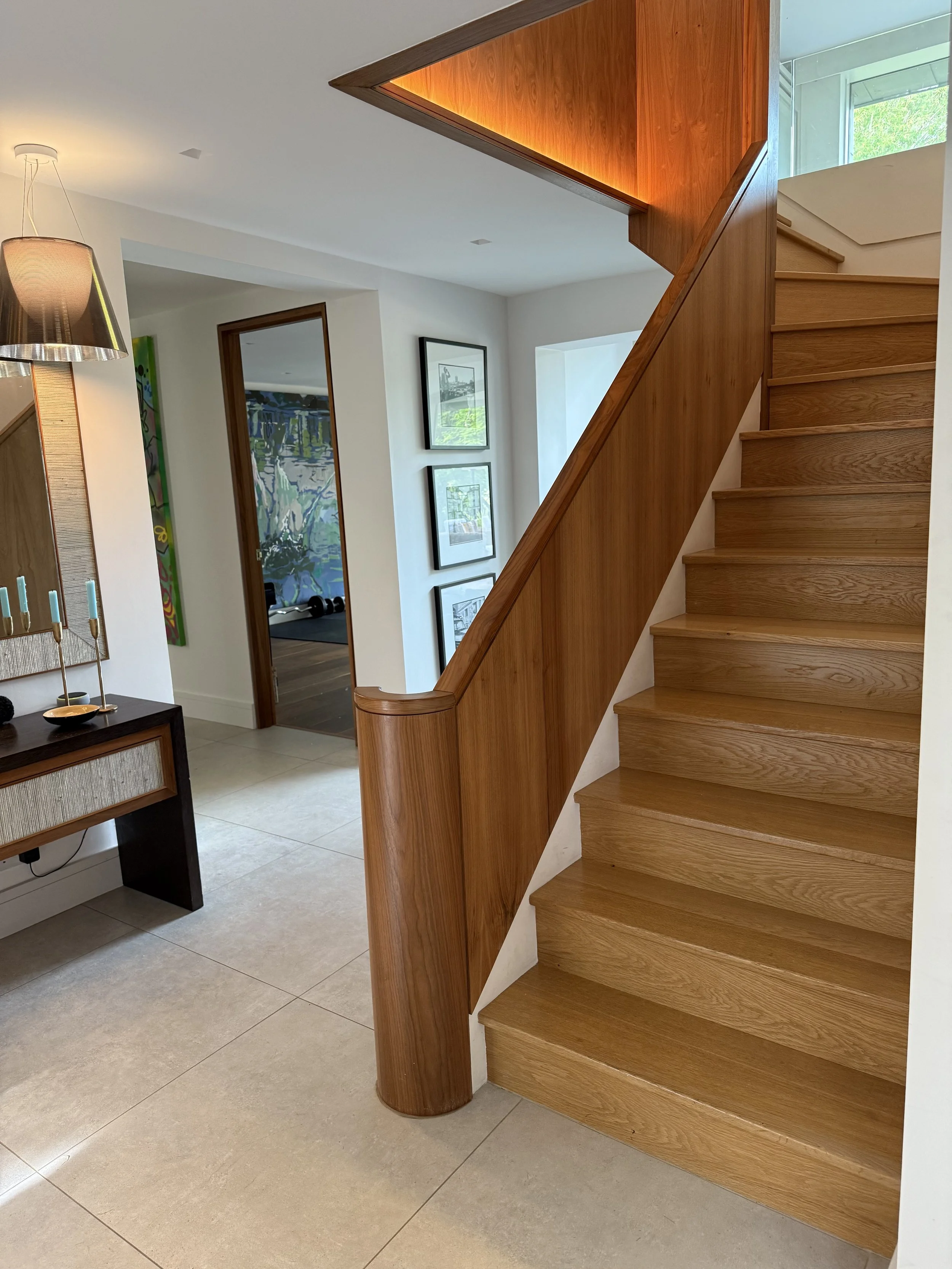Interior view of a modern staircase with wood steps and wooden handrail, leading upstairs in a house with white walls and tiled flooring, decorated with artwork on the walls.