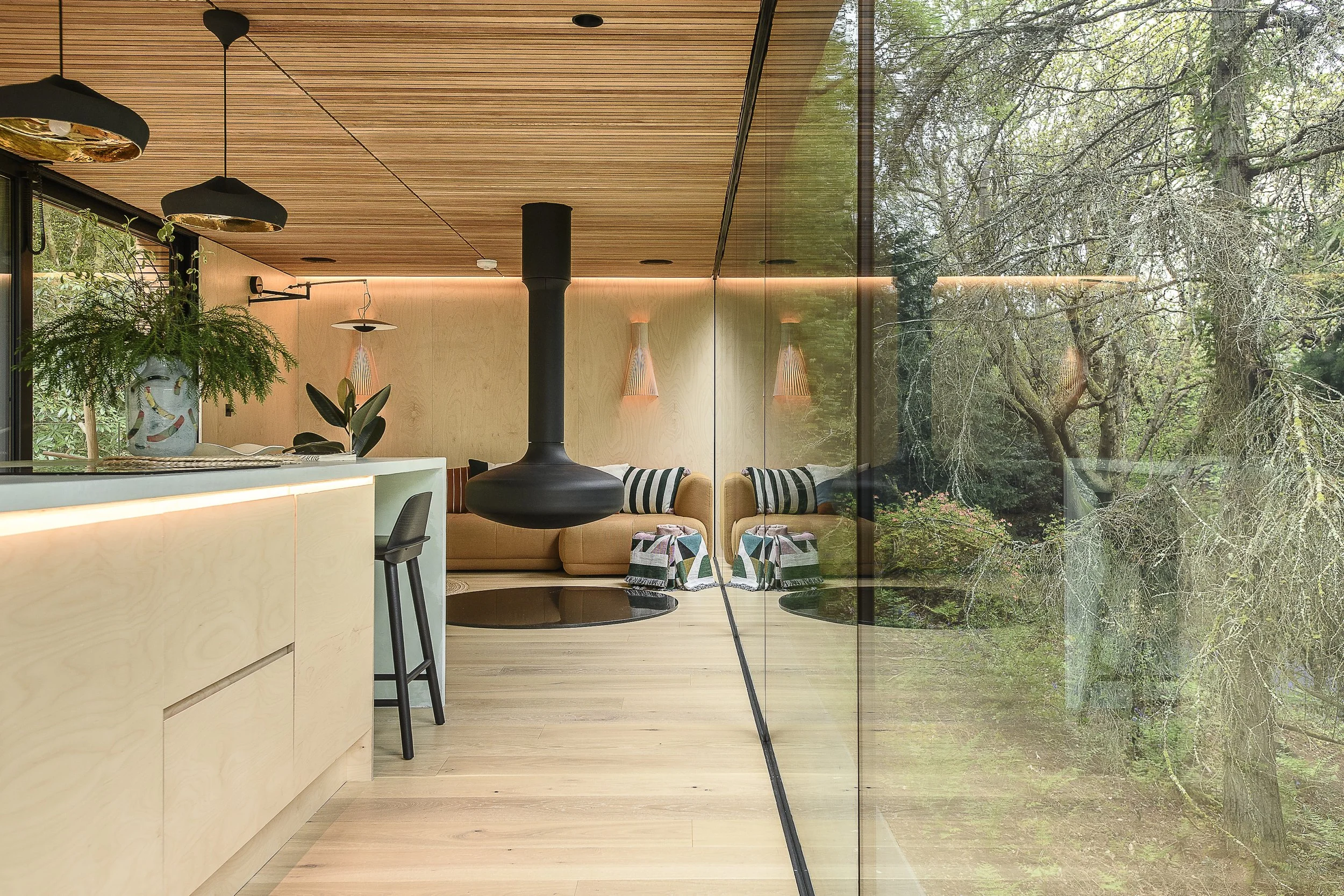 Modern living room with a large glass wall showing trees outside, beige sofa with black and white striped pillows, black ceiling-mounted fireplace, wooden ceiling and floor, (decorative elements like hanging lamps and plants visible).