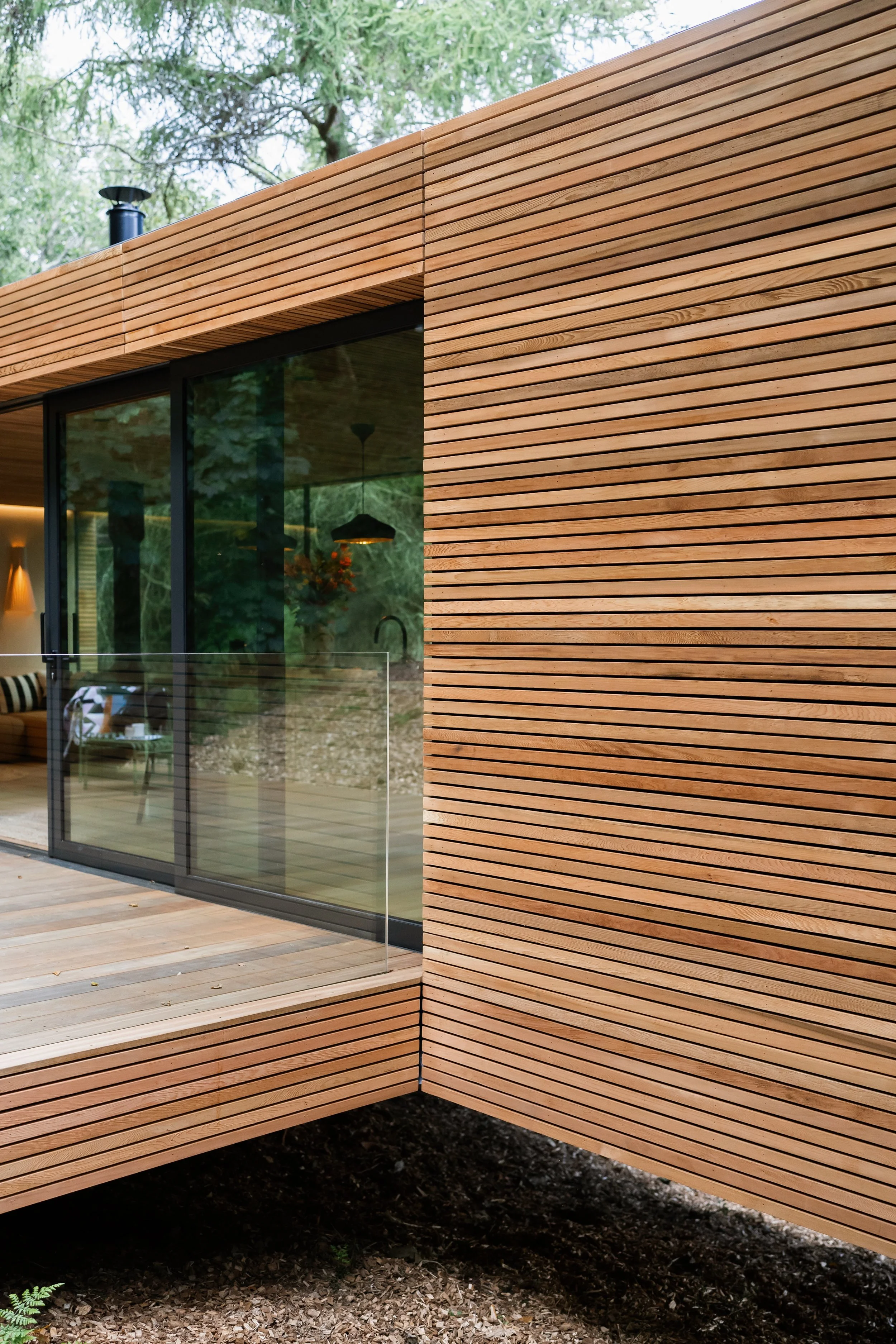 Close-up of modern building exterior with horizontal wooden siding and glass sliding door, showing an interior with a hanging light fixture and furniture.